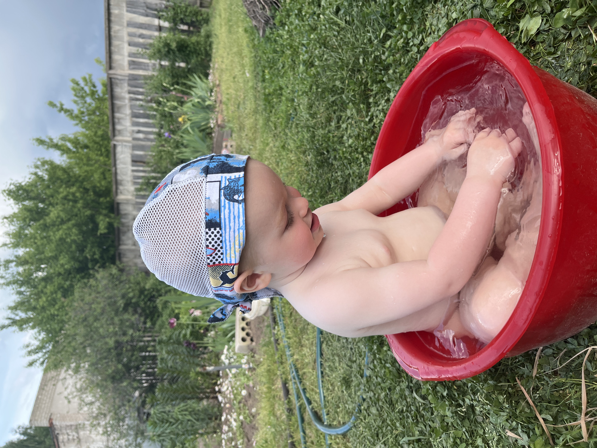 A young child is sitting in a red plastic tub filled with water outdoors. The child is wearing a light-colored cap with a blue and white patterned brim and a white mesh top. The child's body is submerged in the water up to the chest, and their hands are visible, submerged in the water. The tub is sitting on a patch of green grass. In the background, there is a wooden fence, trees, and various plants. The location of Chaikovsky, Russia cannot be determined from this image.