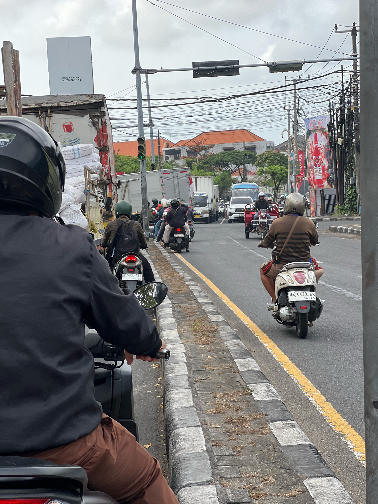 The image depicts a street scene in Denpasar, Indonesia, likely during daytime given the lighting conditions. The primary focus is on traffic, predominantly motorcycles, moving along a paved road. A person wearing a black helmet and a black jacket is in the foreground, seated on a motorcycle, looking ahead. Several other motorcyclists, some with helmets and backpacks, are visible further down the road, participating in the traffic flow.
In the background, large trucks are present, suggesting a commercial or logistics area. One truck is loaded with white sacks. Overhead, a complex network of electrical wires is strung across utility poles, a common sight in urban Indonesian settings. Billboards and signage are also visible, including one with the word "ATLAS" prominently displayed. Traffic signals are present, with one showing a green light. The road has a distinct yellow line marking and a grey and white striped curb. The sky is overcast, indicating a cloudy day. Buildings with orange tile roofs can be seen in the distance, interspersed with trees. The overall impression is of a busy urban street with active traffic.