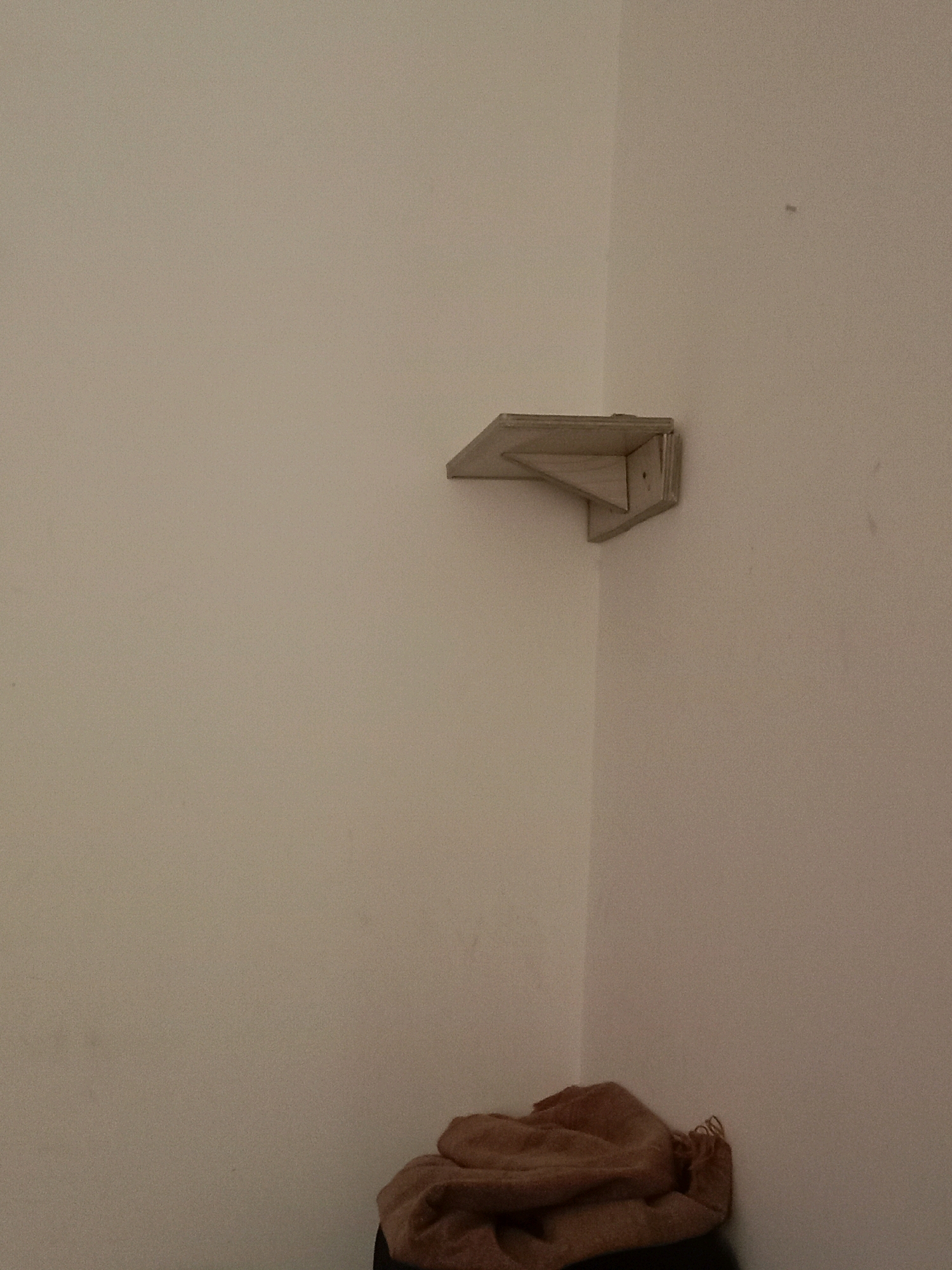 An interior scene depicts a plain, light-colored wall in the corner of a room, likely in Chak Shahzad, Pakistan. Mounted in the corner, approximately eye-level, is a small, triangular wooden shelf. Below the shelf, in the lower right portion of the frame, a pile of folded, light brown fabric, possibly a blanket or towel, rests on an unseen surface. The lighting is dim, casting subtle shadows. There are no people visible in the image.