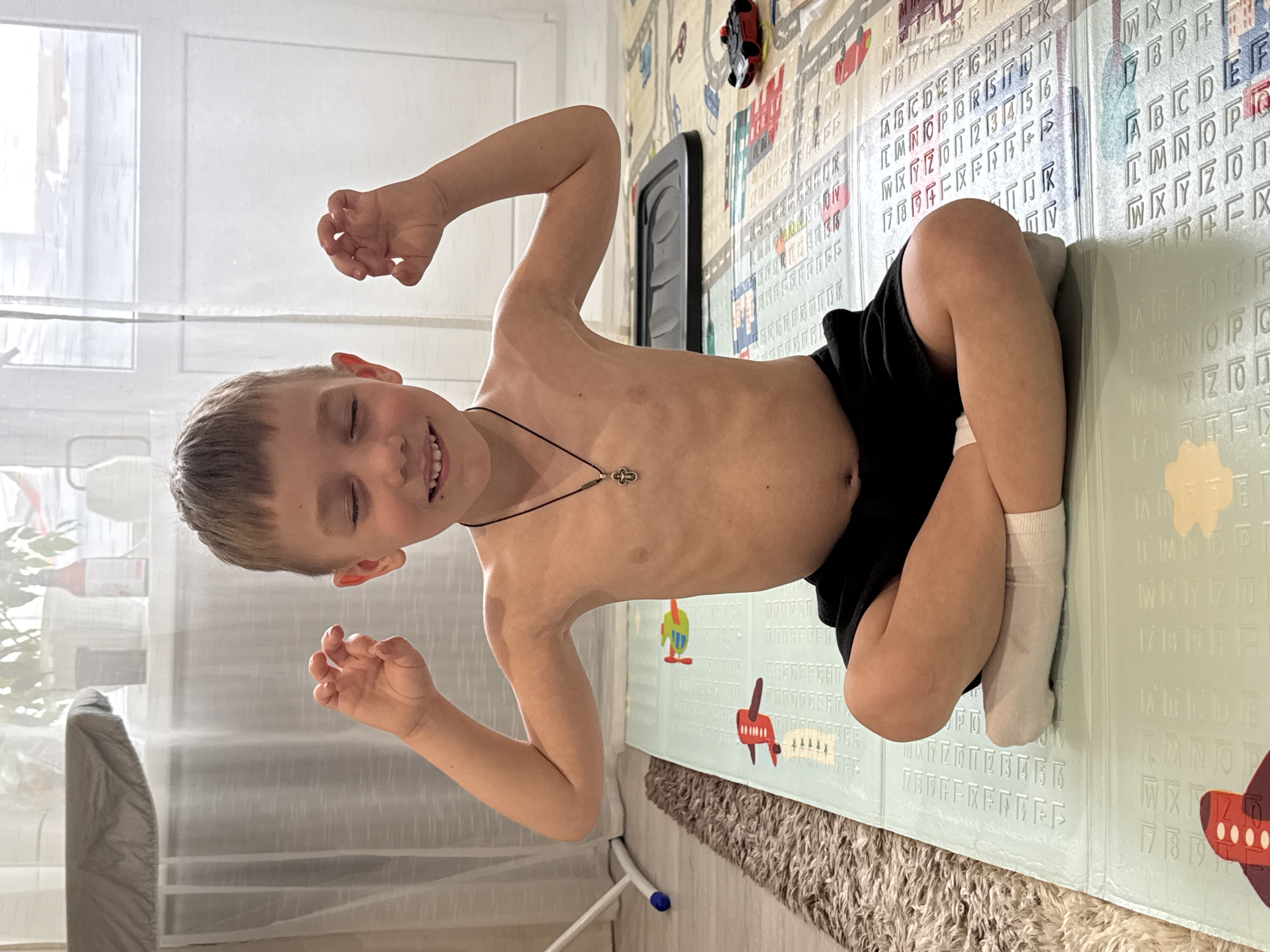 A young boy is lying on his side on a patterned floor mat in a room in Chaikovsky, Russia. The boy has short, light brown hair and is wearing black shorts and white socks. He is shirtless and wearing a necklace with a cross pendant. His eyes are closed and he is smiling. He is holding his hands up near his face. The floor mat has a light blue background and is decorated with letters, numbers, and cartoon vehicles. There is a white wall and window with sheer curtains behind the boy. A carpet is partially visible beneath the edge of the floor mat. A black object is mounted on the wall behind the boy.