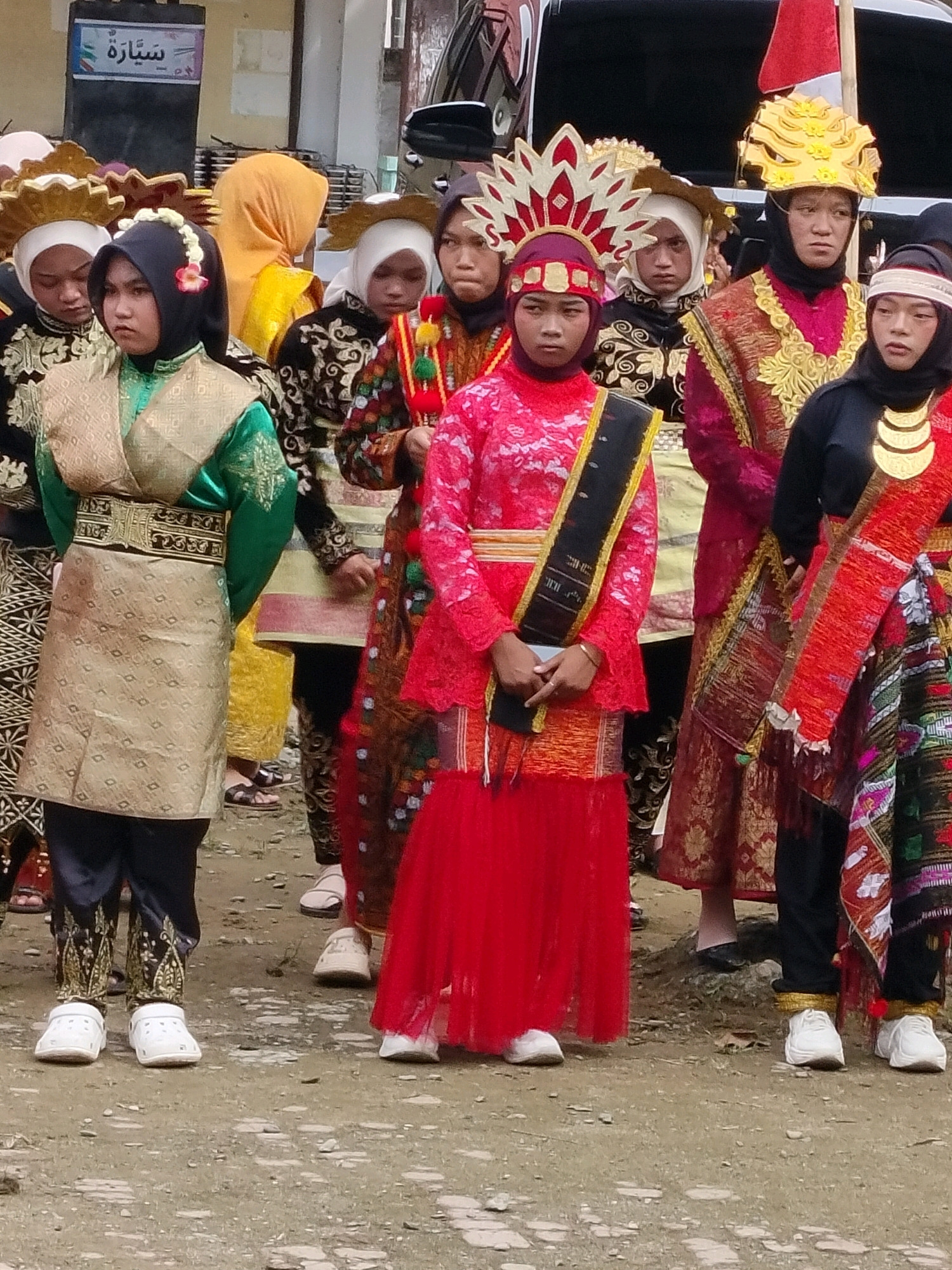 In the city of Purwodadi, Indonesia, a group of young women dressed in traditional attire stand in a line. The woman in the foreground on the left wears a green and gold ensemble, complete with a headscarf adorned with flowers. To her right, another woman is clad in a vibrant red dress, a black sash with gold trim draped across her chest, and a headscarf. The woman to her right is attired in a maroon dress with gold accents and a golden headdress. The woman on the far right wears a black top and a red skirt with a patterned sash. The ground beneath them is a mixture of dirt and gravel. In the background, a vehicle and a building are partially visible.