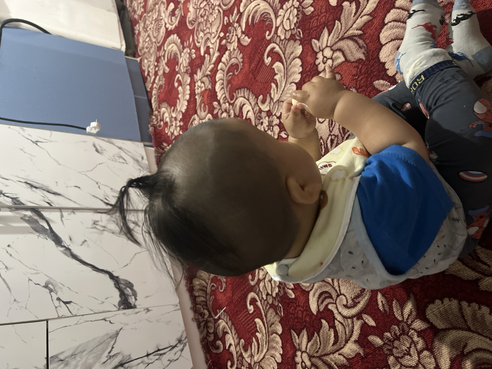 A young child with a small tuft of dark hair is sitting on a red patterned carpet, facing away from the camera. The child is wearing a bib and a blue shirt, with dark pants that have cartoon characters on them. One of the child's feet is visible, wearing a light-colored sock with red and black dinosaur figures. The background includes a wall with a marble pattern and a blue cabinet. The scene appears to be indoors, possibly in a residential setting in Kara-Dıykan, Kyrgyzstan.