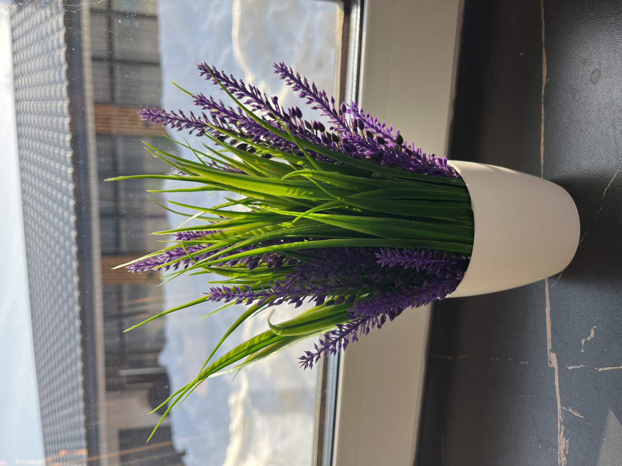 The scene captures an artificial potted plant on a dark, veined surface, likely a windowsill, in Новая Чернушка, Russia. The plant consists of vibrant green, grass-like foliage interspersed with clusters of purple, lavender-like flowers, contained within a simple white cylindrical pot. Beyond a white window frame, an exterior winter landscape is visible, characterized by significant snow cover on the ground. In the mid-ground, a building with a dark-tiled roof and multiple windows, possibly reflecting the sky, stands under a bright, partly cloudy sky. Direct sunlight illuminates the scene from outside the window, creating highlights on the plant and reflections on the glass.