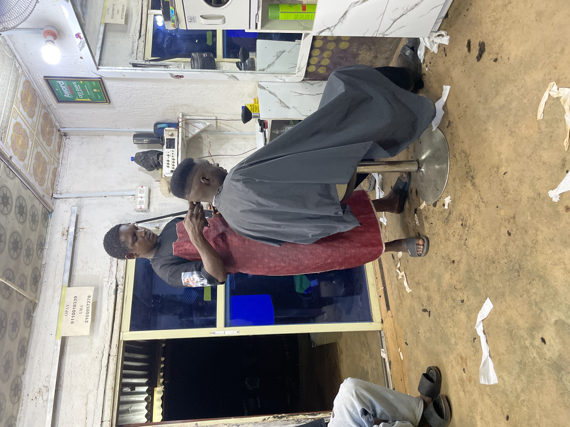 In Durumi, Nigeria, a barbershop scene unfolds with a barber in a red apron and black t-shirt, possibly mid-haircut on a client seated on a stool. The client, draped in a gray cape, is viewed from the back, his face obscured. The barbershop's interior is utilitarian, with a concrete floor littered with snippets of hair and paper. A yellow sign with "OPAY" and account numbers is affixed to the wall above a doorway with a blue-tinted glass panel. To the right, a mirror reflects the interior, showing shelves with various items including cleaning supplies and what appears to be a washing machine. A framed award, possibly for "CEO DAN'S COLLECTION," hangs on the wall. Another person, likely a customer, sits on a bench to the left, dressed in light blue jeans and black sandals. The overall ambiance is that of a bustling, working barbershop.