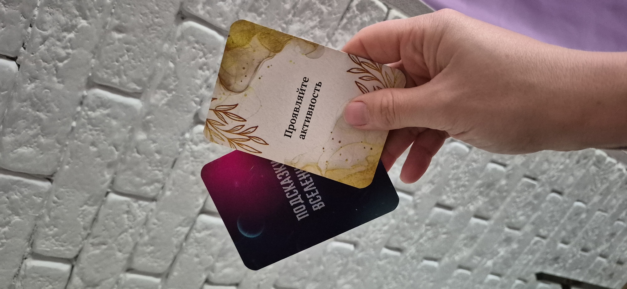 A person is holding two cards against a white brick background. The top card is primarily light-colored with gold accents and text in Russian that translates to "Show activity." The bottom card is darker, featuring shades of black, purple, and blue, also with Russian text and an image of a planet. The location of the scene is unknown.