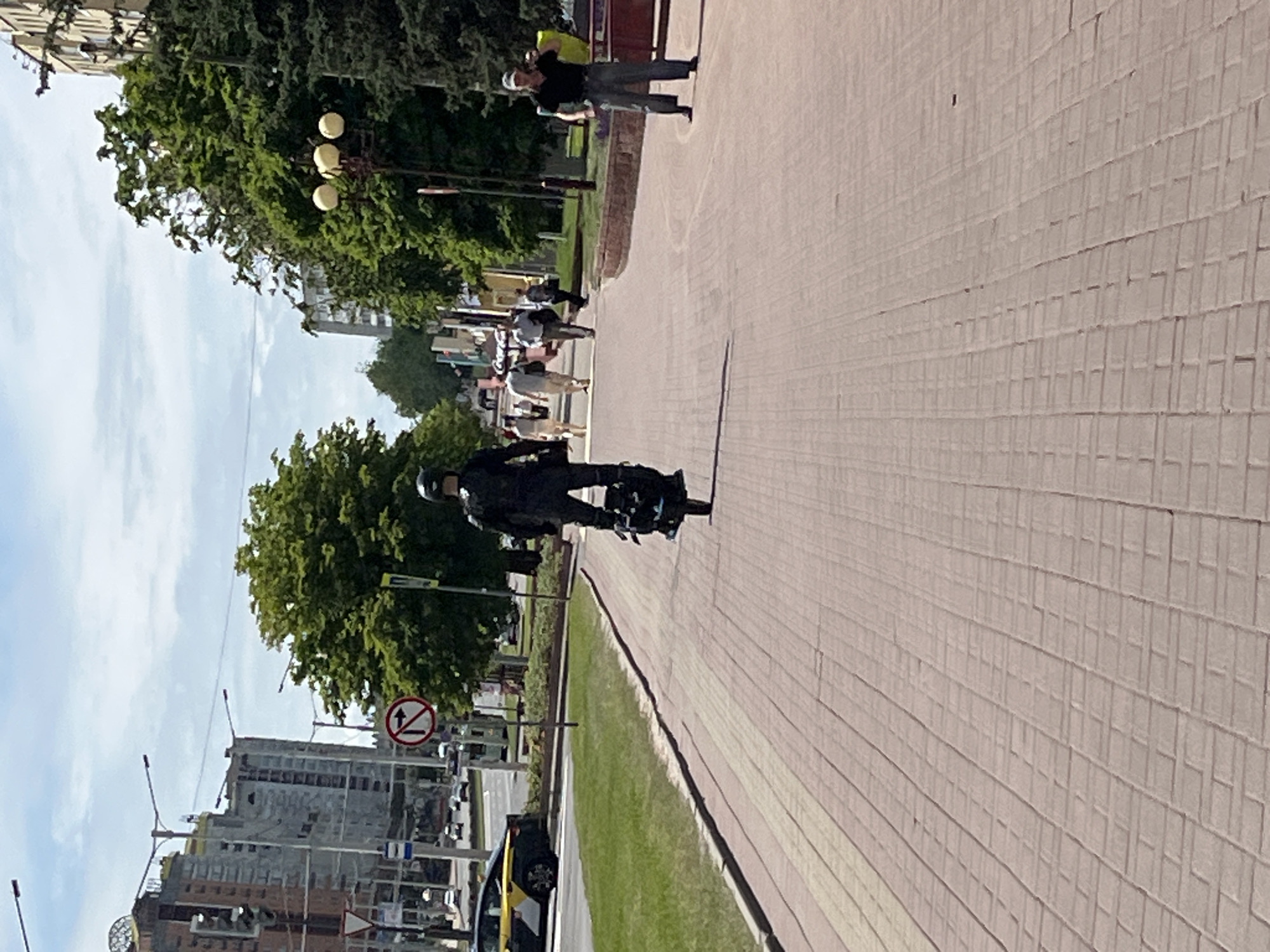 This image depicts a sunny urban scene on a paved pedestrian path in Minsk, Belarus, identifiable by the distinctive spherical structure atop the "House of Football" building visible in the background on the left.
In the foreground, a reddish-brown brick-patterned path, arranged in a stretcher bond, extends towards the background. Shadows from an unseen object are cast diagonally across the path from the left.
In the midground, a person wearing dark clothing and a helmet is riding an electric unicycle, viewed from the rear, traveling away from the camera. The unicycle is dark-colored with visible blue accents. To the left of the path, a strip of green grass separates it from a multi-lane asphalt road where a yellow and black taxi is visible, partially obscured, near what appears to be an intersection. A red and white "No Right Turn" sign is prominently displayed on the left side of the road, along with other traffic and informational signs.
Further along the pedestrian path, several pedestrians are walking away from the viewer. On the far right, a man wearing a black polo shirt, grey pants, and a light-colored baseball cap stands facing the viewer with his arms slightly raised. The right side of the path is bordered by mature green trees, among which are visible streetlights with multiple globe fixtures.
The background on the left features several multi-story buildings, including the aforementioned "House of Football" with its prominent golden sphere. The sky above is light blue with scattered white clouds. The right side of the background shows more dense foliage and trees with additional buildings visible through them, characteristic of a city environment.