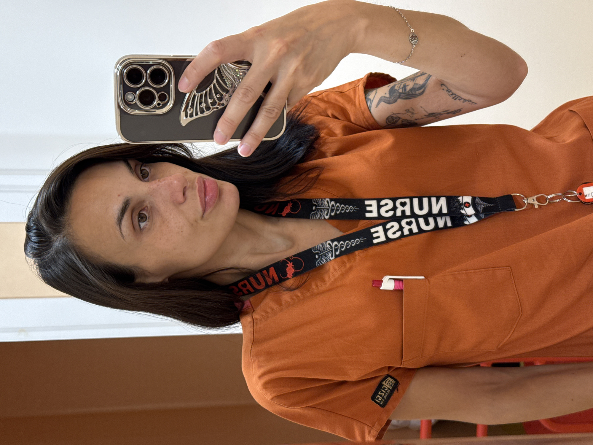 A woman with dark hair and freckles takes a selfie in a mirror. She is wearing an orange scrub top with a lanyard around her neck. The lanyard has medical symbols and the word "NURSE" printed on it. She has a pen in the chest pocket of her scrub top. She is also wearing a silver bracelet on her right wrist and has tattoos on her left arm. The background is a plain wall and a doorway. The scene appears to be indoors, possibly in a hospital or clinic setting.