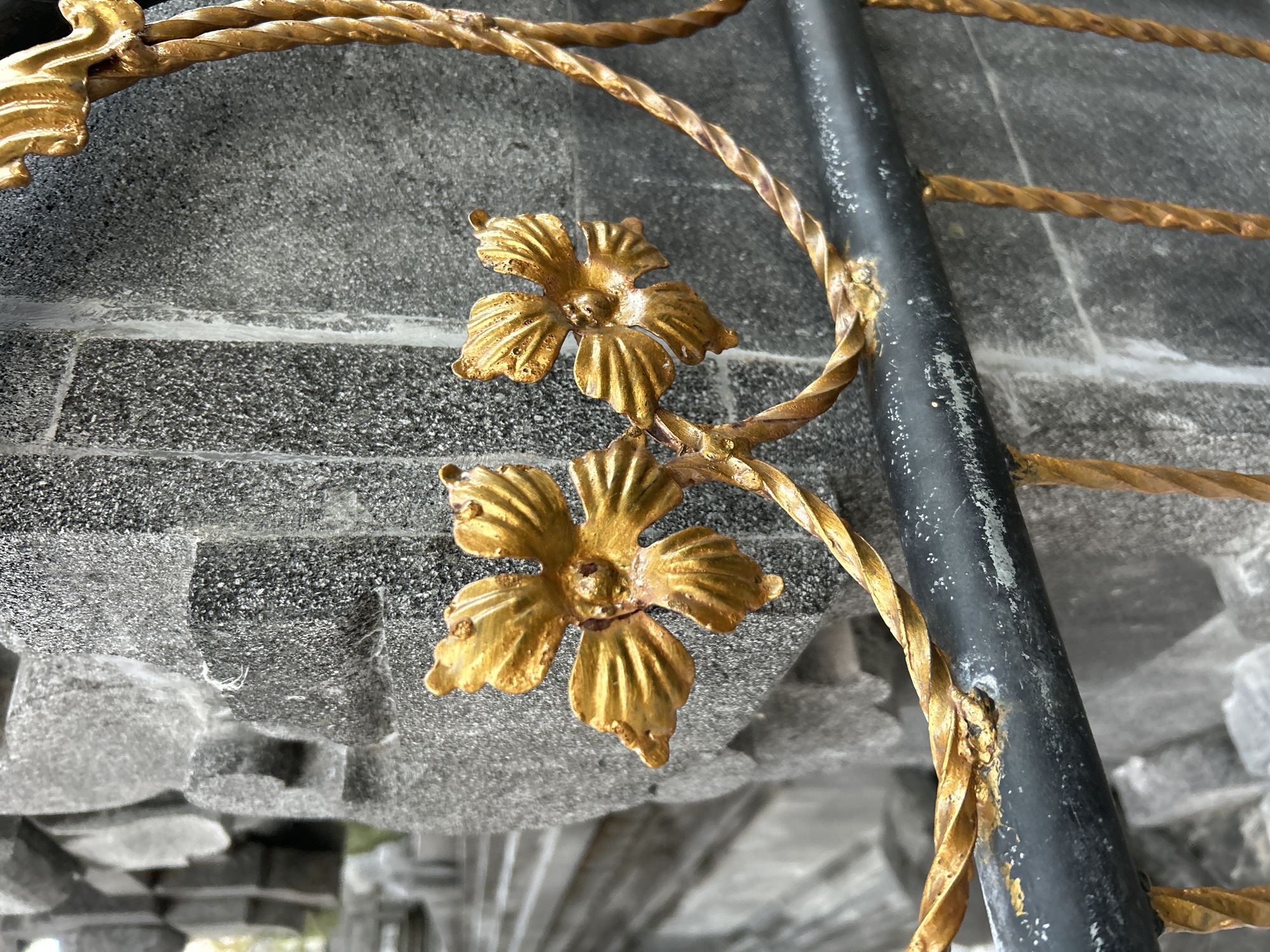 In Abiansemal, Indonesia, a close-up shot focuses on a black metal railing adorned with ornate golden decorative elements resembling flowers and twisted vines. Behind the railing, a textured grey stone wall serves as the backdrop, with further stone structures visible in the blurred background. The lighting is natural, and the scene appears to be outdoors.