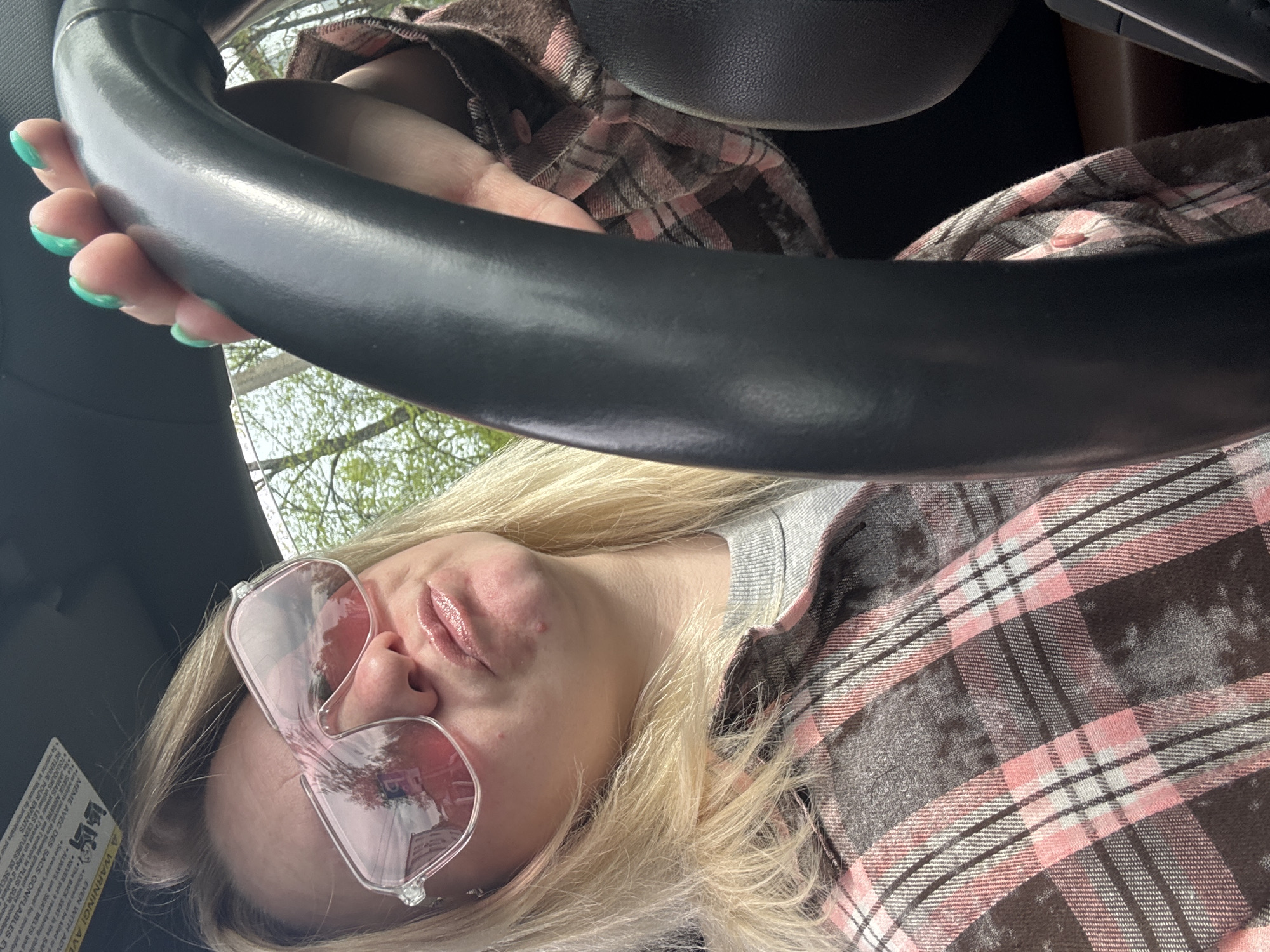 A selfie from inside a car. A blond woman with pink glasses holds the steering wheel with her left hand. She wears a pink and gray plaid shirt. The car's dashboard and a warning label are visible. The location is not discernable from the image.