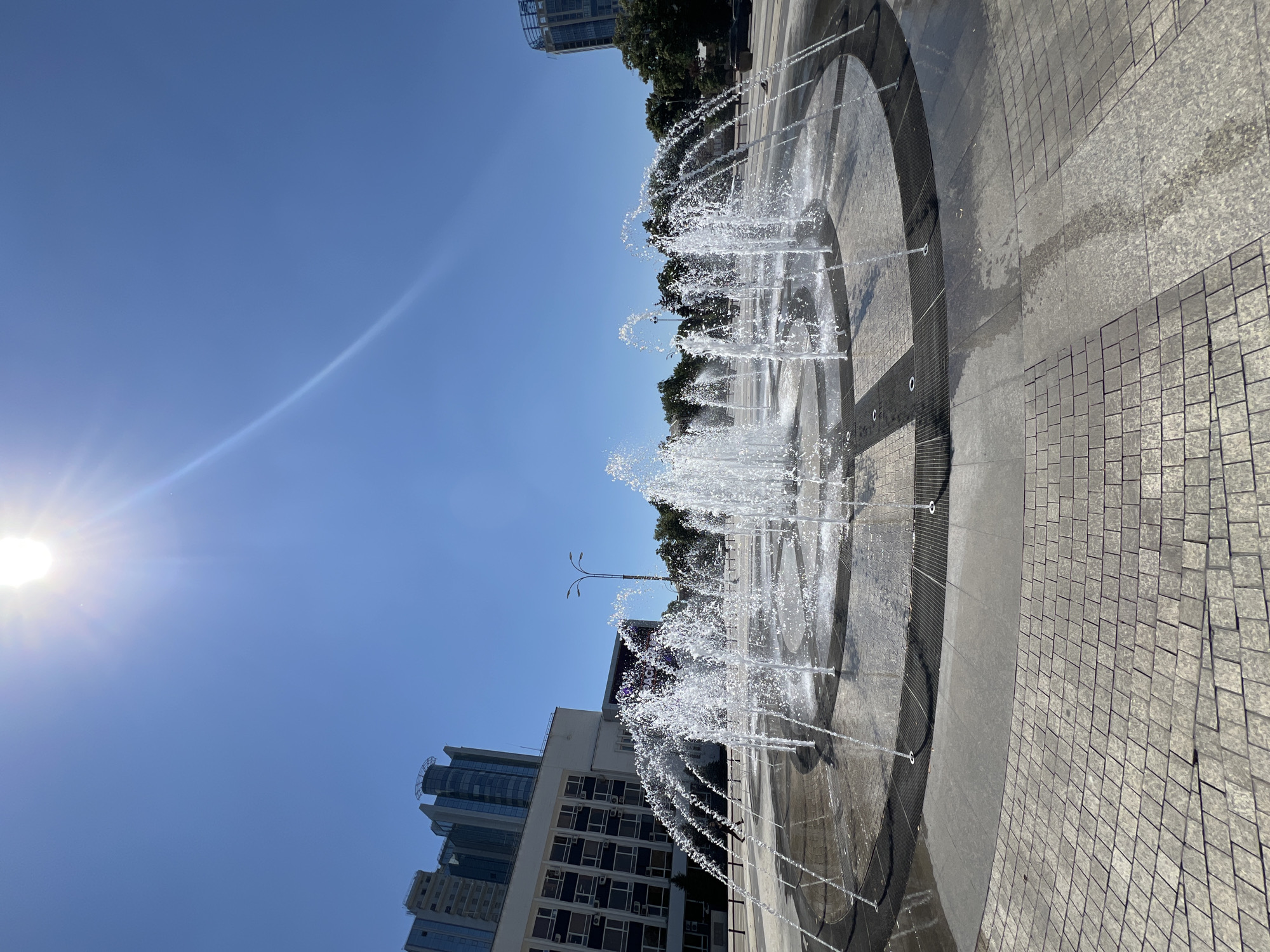 This is a low-angle, wide shot of a modern public plaza in Krasnodar, Russia, featuring a circular fountain with numerous water jets spraying upwards in a synchronized, arching pattern. The fountain is situated on a paved surface with concentric circles of dark gray and light gray stone tiles, bordered by a darker ring with metal grates.
In the background, several modern, multi-story buildings are visible under a clear, bright blue sky. The building to the left is a cream-colored structure with a grid of dark windows and a darker, glass-fronted section on top. To its right are taller, contemporary glass skyscrapers. Further to the right, another glass-fronted building with a curved roofline is visible.
Trees line the perimeter of the plaza behind the fountain, and streetlights are present. The sun is high in the sky, causing lens flare and casting bright highlights on the water jets and pavement. A single person is walking across the plaza in the middle ground, to the left of the fountain. A pigeon is also visible near the edge of the plaza on the right. The overall atmosphere is one of a sunny, urban public space.