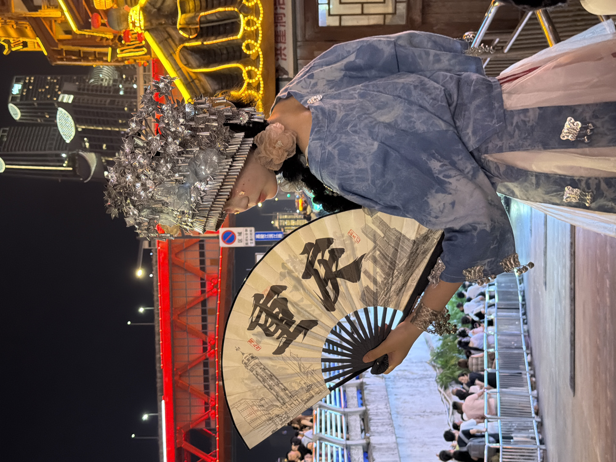 The image captures a person dressed in traditional attire, holding a fan, at an event or performance likely set in the Yuzhong District of China. The subject is wearing a blue, tie-dye patterned robe adorned with ornate silver clasps and cuffs. A striking, intricate silver headdress with floral embellishments sits atop their head. A delicate pink flower is pinned to their hair near their ear.
The fan is a prominent object, open and displaying a black ink painting of what appears to be a cityscape with a prominent tower, possibly a landmark. Two large Chinese characters are painted in bold black strokes on the fan.
The background suggests an evening setting, with a vibrant display of yellow and red lights from what could be a building or a stage setup. In the distance, modern skyscrapers are visible, indicating an urban environment. A red, illuminated structure, possibly a bridge or walkway, is also a significant element in the scene.
Below the subject, a crowd of people can be seen, seated or standing behind a silver railing, suggesting they are spectators at an event. The overall atmosphere appears festive and engaging, possibly a cultural celebration or a performance. The lighting and attire suggest the event is taking place at night. There are no discernible weather cues.