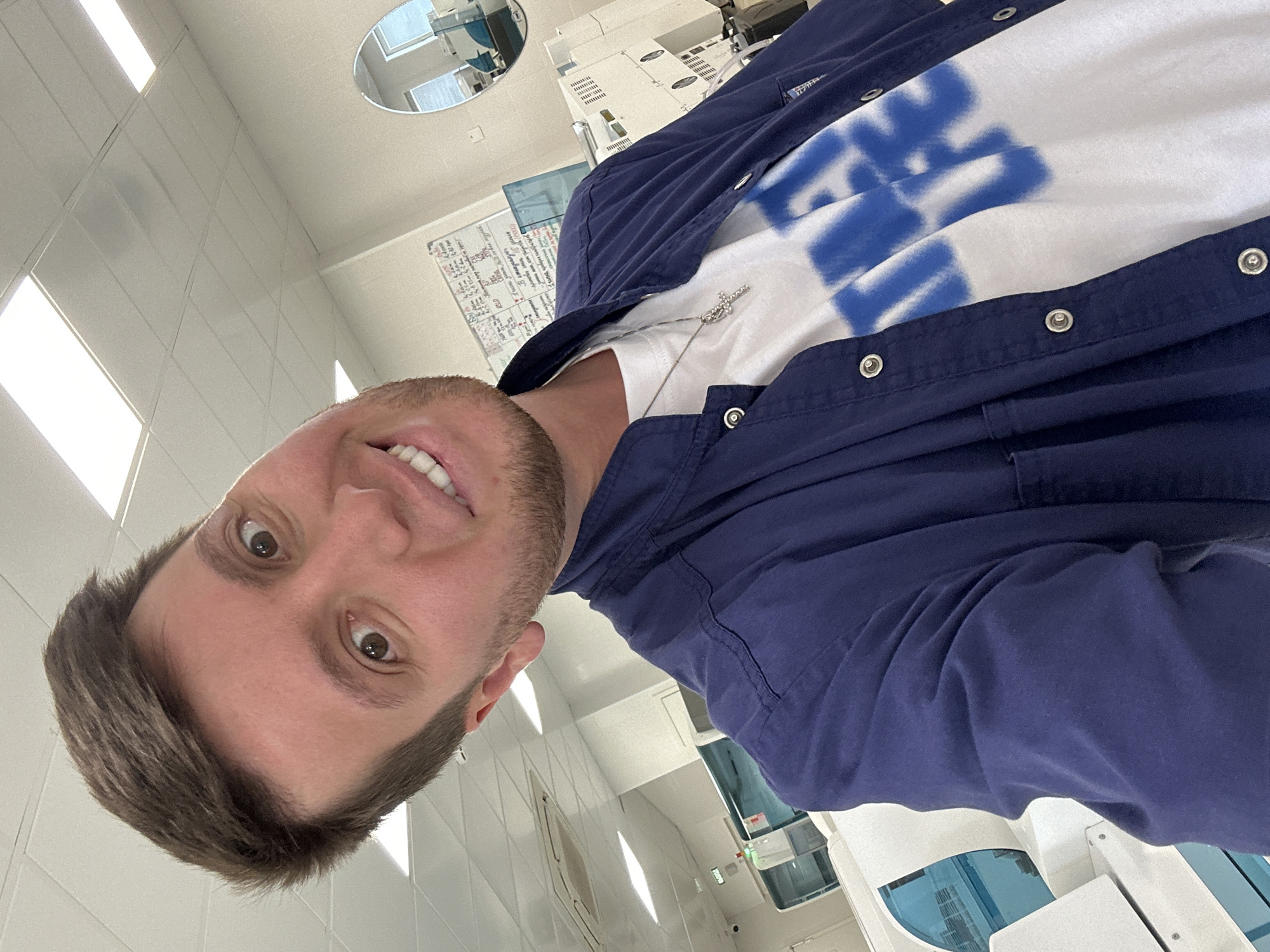 The media file is a selfie taken by a person indoors, likely in a laboratory or medical facility given the equipment visible in the background. The subject is a man with short brown hair and a beard, smiling with his mouth open, revealing his teeth. He is wearing a white t-shirt with a blue graffiti-style design and a navy blue jacket over it. A silver necklace with a cross pendant is visible. The background shows a tiled ceiling with fluorescent lighting, and in the distance, there are various pieces of white laboratory equipment with blue accents. There is also a circular mirror on the ceiling and a whiteboard with handwritten text. The overall lighting suggests it is daytime. The image is rotated, with the subject's head at the bottom and his feet (implied) at the top. The setting appears to be sterile and functional, common in scientific or healthcare environments. The subject's expression conveys a sense of happiness or amusement. No specific time of day or weather can be determined from the interior setting, but the bright lighting suggests it is not nighttime. There is a whiteboard with some illegible text, likely notes or instructions related to the facility. The location context is stated as Moscow, Russia.