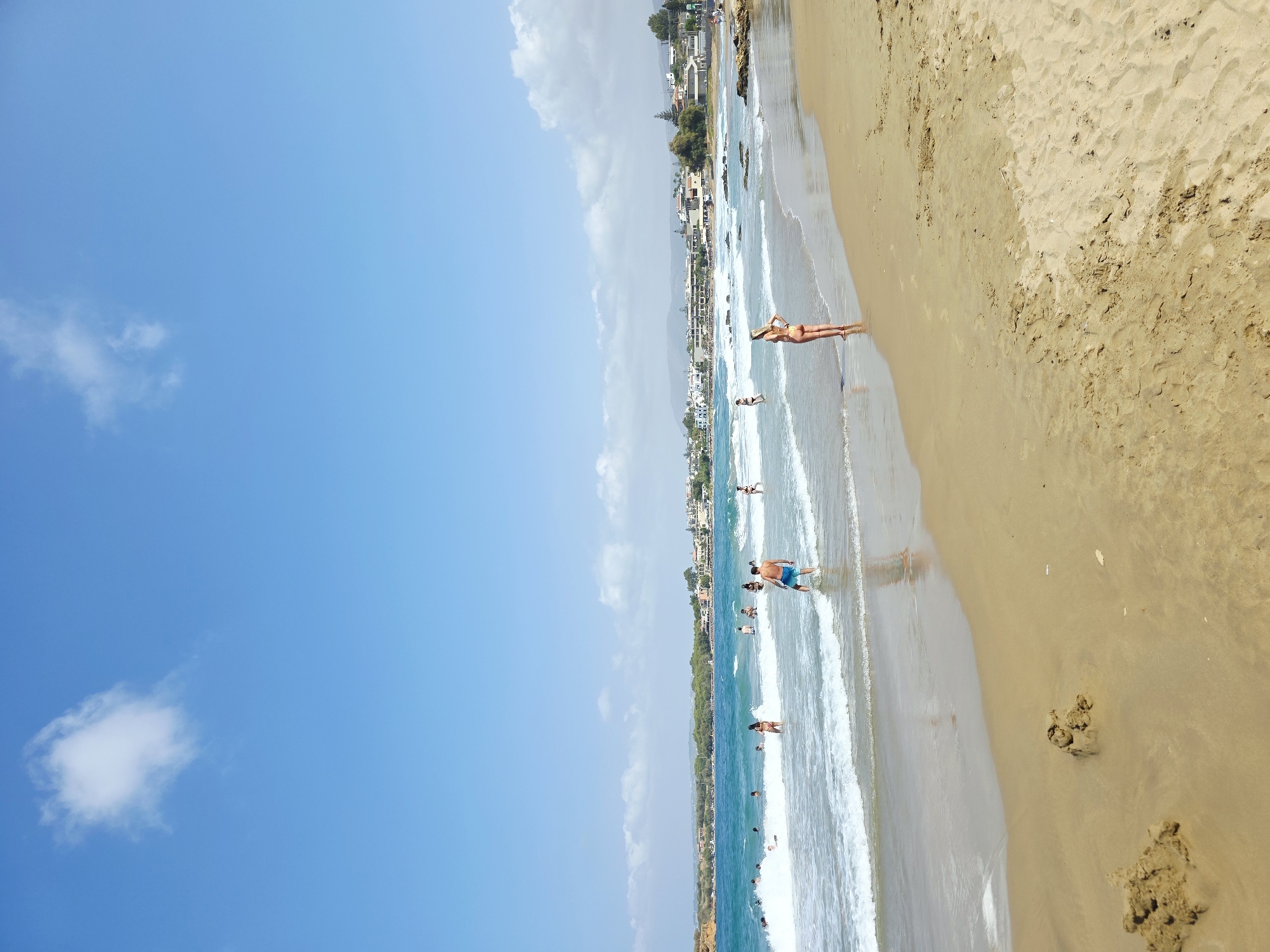 The image depicts a sunny day at a beach in Chania, Greece. The foreground is a sandy beach with a few piles of sand. In the middle ground, people are swimming and wading in the turquoise water, with waves breaking on the shore. A woman in a bikini stands at the edge of the water, facing away from the camera. Further in the distance, along the coastline, are buildings with white walls and terracotta roofs, characteristic of Greek architecture. Lush green trees are interspersed among the buildings. The sky is a vibrant blue with a few scattered white clouds.