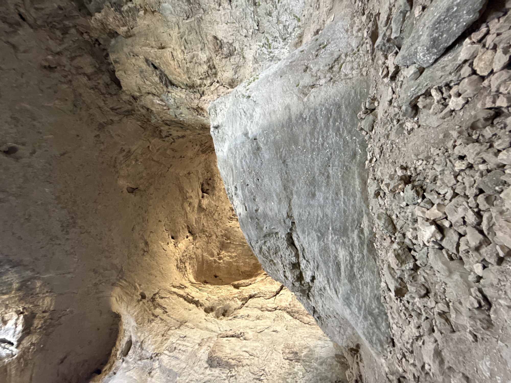 The image captures the interior of a cave in Karlukovo, Bulgaria, likely the Prohodna Cave, also known as the "Eyes of God" cave. A large, irregularly shaped boulder dominates the foreground, appearing to be made of grey rock. The boulder's surface is textured with various pits and indentations. Surrounding the boulder is a dense collection of smaller rocks and debris, suggesting a natural, undeveloped environment within the cave. The cave walls rise around the boulder, exhibiting rough, uneven textures. Light streams in from an opening higher up in the cave, illuminating parts of the walls and creating shadows. The overall impression is of a natural, geological formation, with the boulder being a significant feature within the cavernous space.