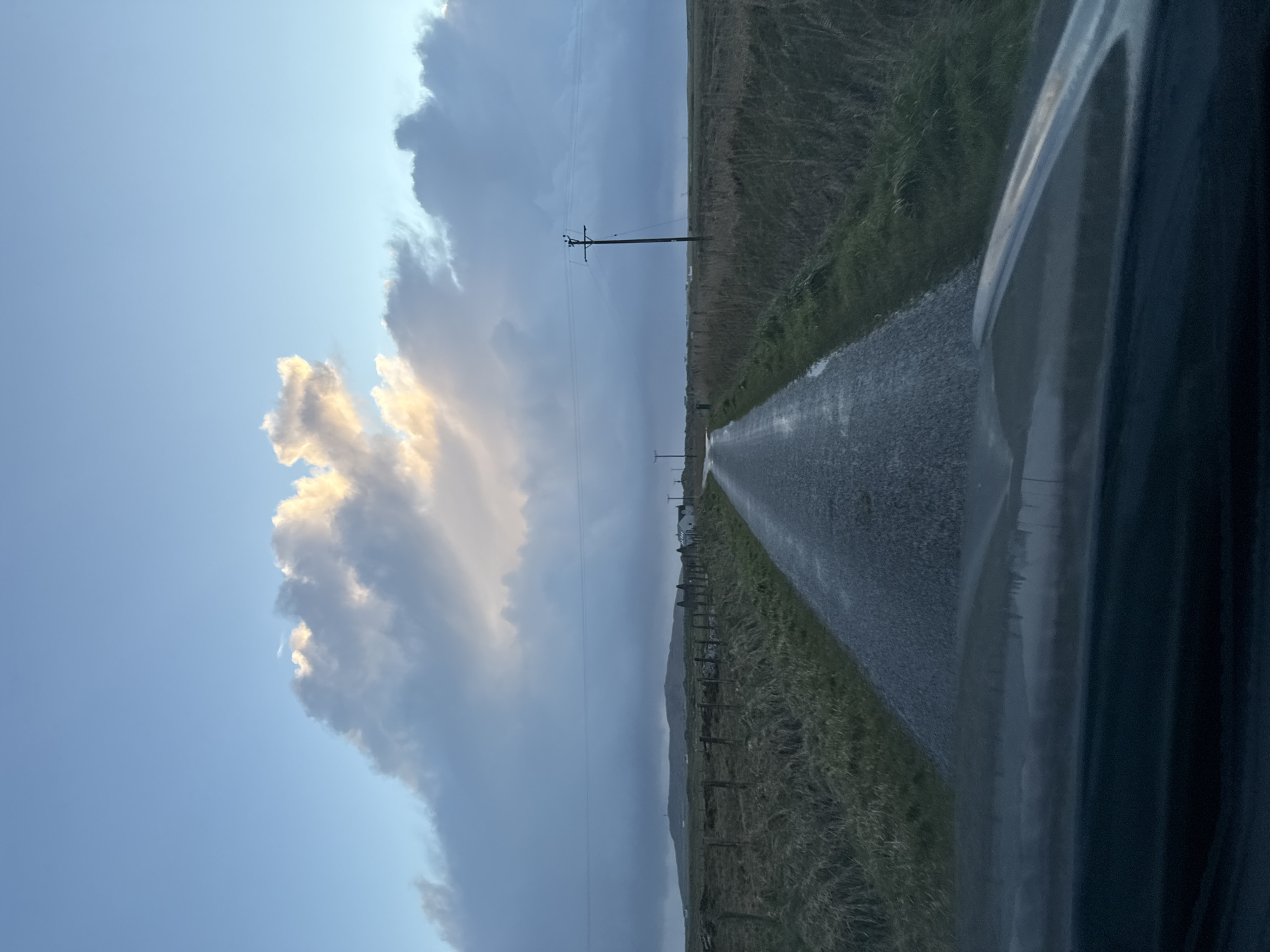 The image shows a road running through a rural landscape. The road is wet, suggesting recent rainfall. The sky is partially cloudy with some patches of blue visible. There are utility poles along the roadside. The vegetation along the road consists of tall grass. A portion of a vehicle is visible in the foreground, suggesting the image was taken from inside a car. The exact location in the city of Unknown, Unknown cannot be determined.