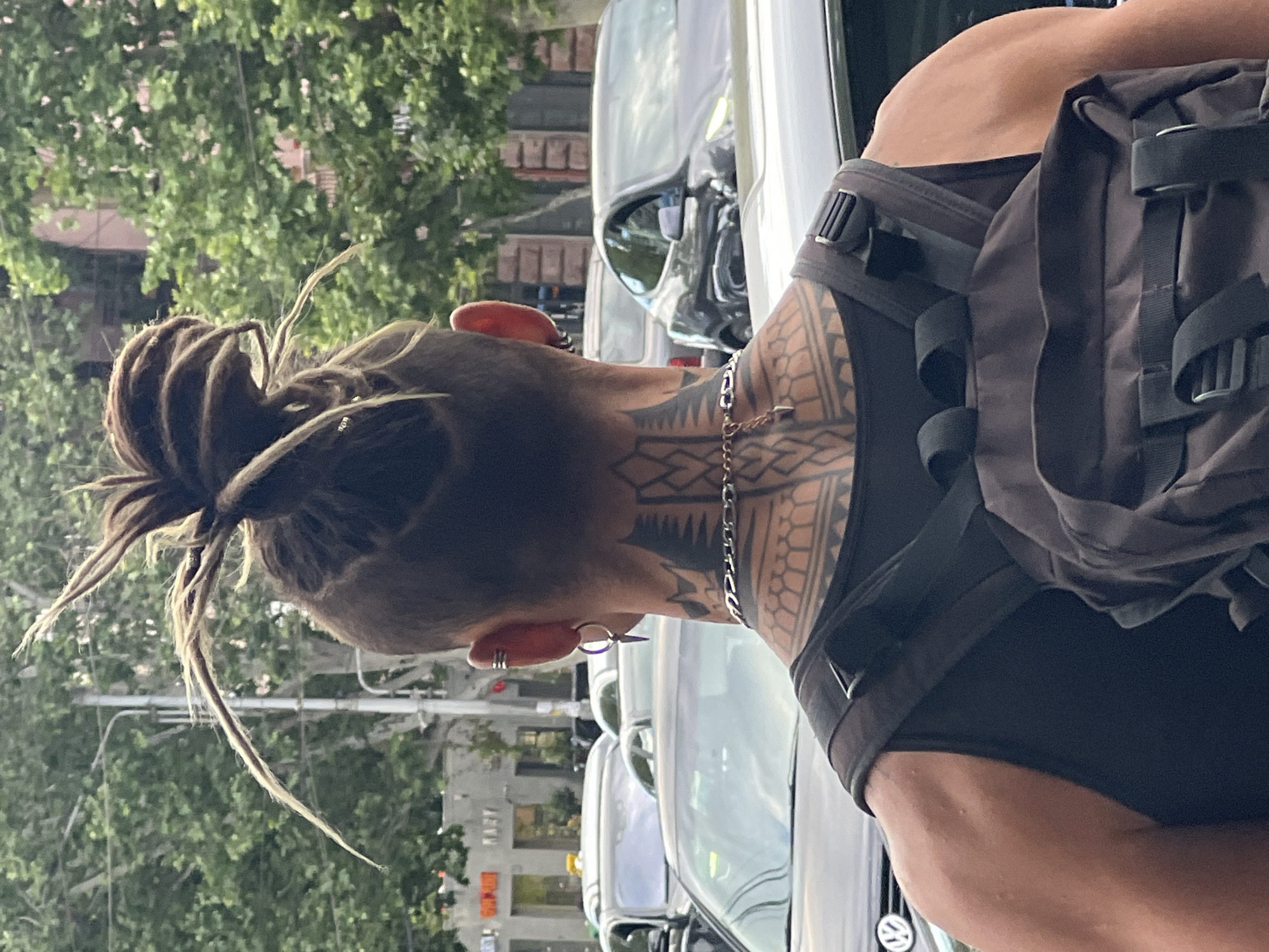 The image captures a person from behind, showing their head, neck, right shoulder, and upper back in an outdoor urban setting, likely in Yerevan, Armenia.
**Subjects:** The central subject is a person with dreadlocks, which are styled into a bun or ponytail, exposing a shaved undercut on the sides and back of their head. Several long, thin dreadlock strands hang loose. The person has multiple ear piercings, including hoops and studs. Extensive geometric or tribal-style tattoos cover the back of their neck, extending down the spine area, and onto their right shoulder and upper arm. They are wearing a dark, possibly black, sleeveless top or tank top, and a dark-colored backpack with visible straps and buckles is worn over it. A silver chain necklace is also visible around their neck, partially obscured by the hair and tattoo.
**Scene and Setting:** The background features green foliage from trees, suggesting a street lined with greenery. Several cars are visible, parked or moving slowly, indicating a city street or roadside. Buildings can be faintly seen through the trees in the distance.
**Activity:** The person appears to be standing or walking away from the viewer, moving through or observing the urban environment. No specific activity beyond presence is discernible.
**Notable Details:**
* **Time of Day and Weather:** The lighting suggests daytime, possibly late morning or afternoon, with diffused light indicating either an overcast sky or dappled sunlight through trees.
* **Tattoos:** The prominent tattoos are a key feature, characterized by intricate geometric patterns that create a strong visual statement on the neck and shoulder.
* **Physique:** The person exhibits a muscular build, particularly in their upper back and shoulders.
* **Accessories:** The backpack and various ear piercings add to the individual's style.
**Visible Text:** There is no legible text visible in the image.