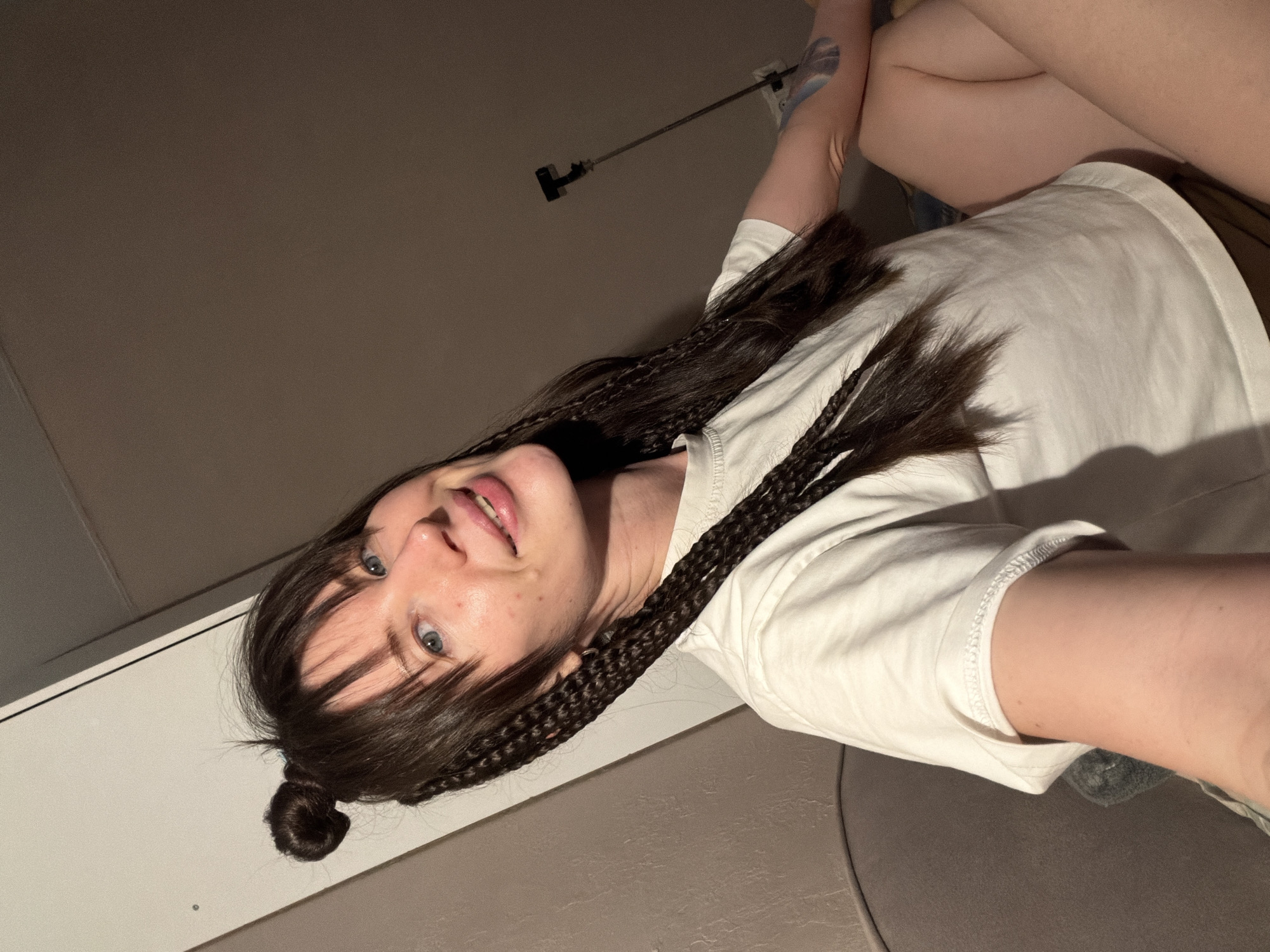 The image is an upward-facing selfie of a young woman, significantly rotated clockwise, suggesting she is lying down or reclining. She has fair skin, light blue eyes, and long, dark hair styled with numerous braids integrated into her hair, along with bangs framing her face. A small bun made of her hair is visible towards the lower left of the frame. She is wearing a plain white t-shirt and displays a gentle smile, with her lips slightly parted and her eyes looking directly at the camera.
Her left arm is visible, and a faded or light blue tattoo can be seen on her upper left arm/shoulder area. Parts of her legs are visible in the upper right, further indicating a reclining posture. The background consists of a plain, light brown or grey wall/ceiling. A thin, dark rod or pole with attachments is visible in the upper left background, possibly a part of the room's fixtures or furniture. The lighting suggests an indoor setting, likely evening or night. The location context is Perm, Russia. No visible text is present.