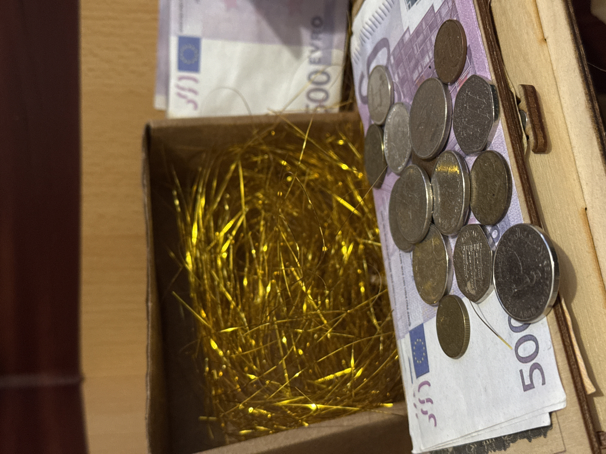 The image presents an overhead view into an open container, featuring a collection of money and golden decorative material. On the right side, a light-colored wooden box is partially visible, showing a simple wooden latch mechanism.
Within this container, a stack of 500 Euro banknotes is prominently displayed, with the "500 EURO" denomination clearly visible. Resting on and around these banknotes is an assortment of various coins. The coins display diverse designs, indicating a mix of international currencies. Visible examples include a British coin featuring the profile of Queen Elizabeth II, an octagonal coin with an indistinct design, and another coin with what appears to be Arabic script, possibly an Omani Rial. Several other coins of varying sizes and metallic finishes, likely Euro coins, are also present.
To the left, a separate section of the container, appearing to be made of brown cardboard, is filled with a dense, shimmering mass of golden tinsel or shredded metallic paper. This material creates a striking contrast with the currency and the wooden elements. The lighting in the image suggests an indoor setting, possibly under artificial light, which accentuates the reflective qualities of the coins and tinsel. No people are visible in the scene. The overall impression is that of a personal collection of money, potentially stored in a decorative or significant manner, such as a treasure chest or savings box.