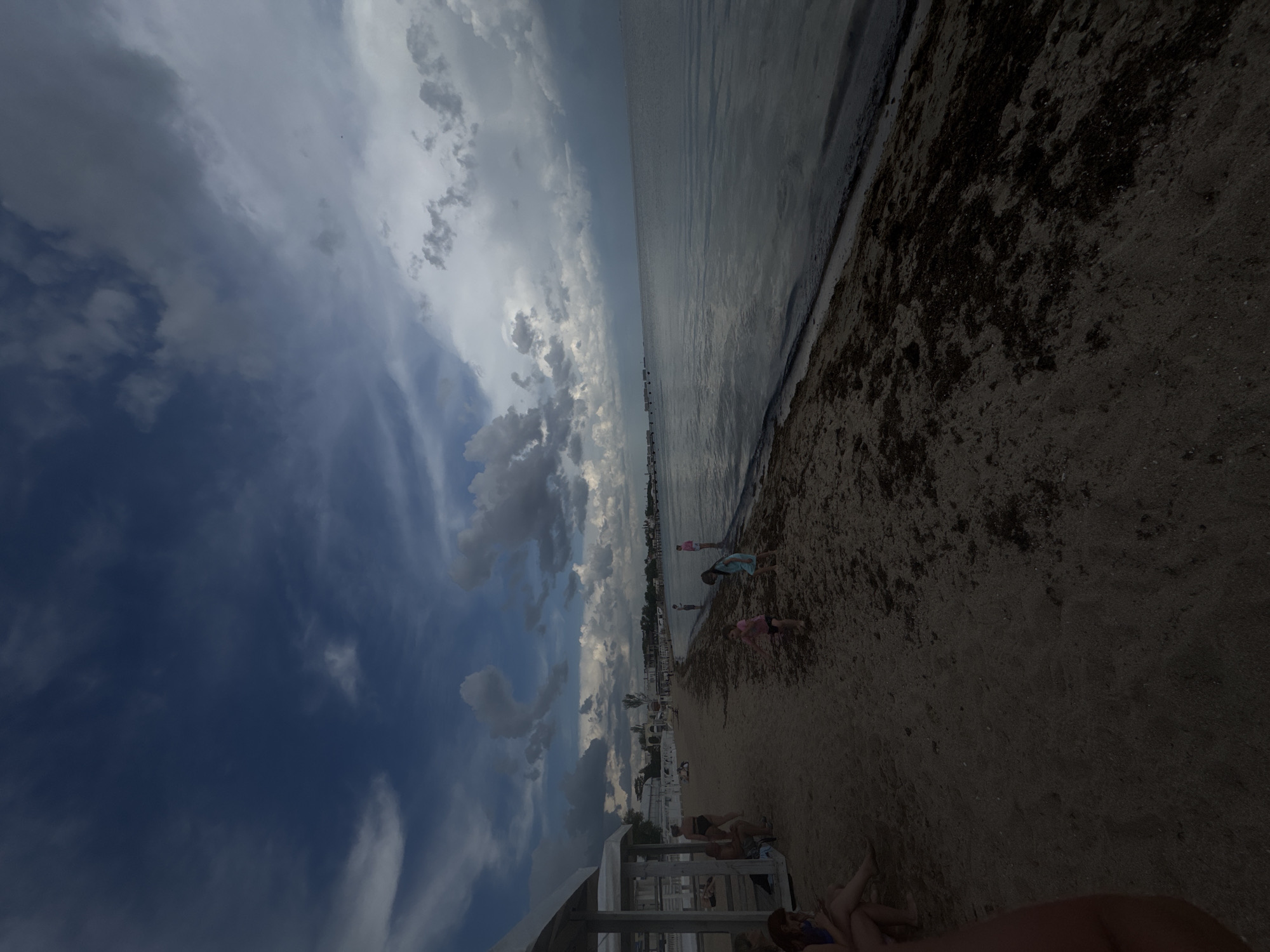 The image depicts a beach scene in Yevpatoriya, Ukraine. The view is oriented with the beach on the right and the sky on the left. The sky is dominated by a mix of dark blue and white clouds. The sea is calm and appears gray. A narrow strip of dark material lines the shoreline, likely seaweed or debris.
Several people are on the beach. Near the shoreline, two children are playing. Further inland, there is a cluster of people relaxing on lounge chairs under the shade of structures.