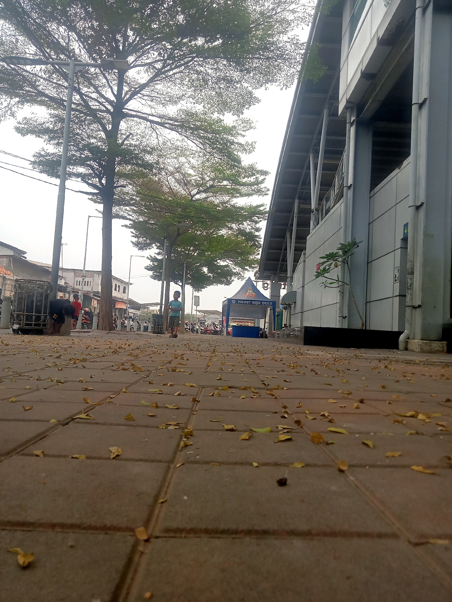 The image depicts a low-angle view of a paved walkway in an urban environment, with scattered dry leaves on the ground. To the left, a metal cage-like structure stands near a tree, with two individuals partially visible behind it. Further down the walkway, a young boy in a blue t-shirt and shorts stands facing the camera. A large, modern building with a covered walkway supported by columns runs along the right side of the scene. In the background, various vehicles, possibly motorcycles, are parked. A blue and red tent with text is visible in the middle distance. The sky is overcast, suggesting diffused lighting. The overall impression is that of a pedestrian area adjacent to a transit hub or a commercial district.