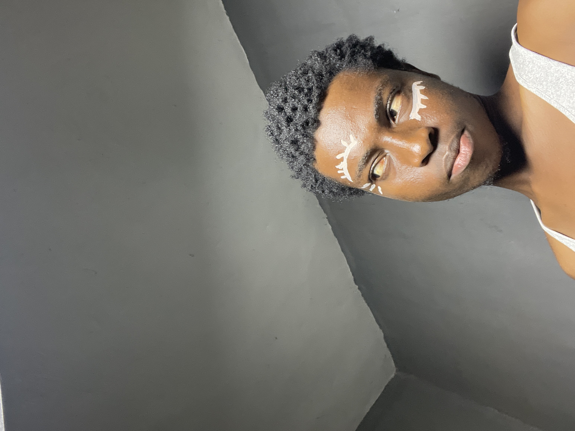 A close-up portrait shows a young person with dark skin and short, curly black hair, wearing a white tank top. They are looking to their left with their eyes slightly narrowed. White face paint, resembling stylized eyelashes or abstract designs, is applied above and below their eyes. The background consists of a dark gray wall meeting a lighter gray ceiling at a sharp angle, creating a stark, dimly lit environment. There is no indication of the specific location within Nkpor, Nigeria, in the image itself.