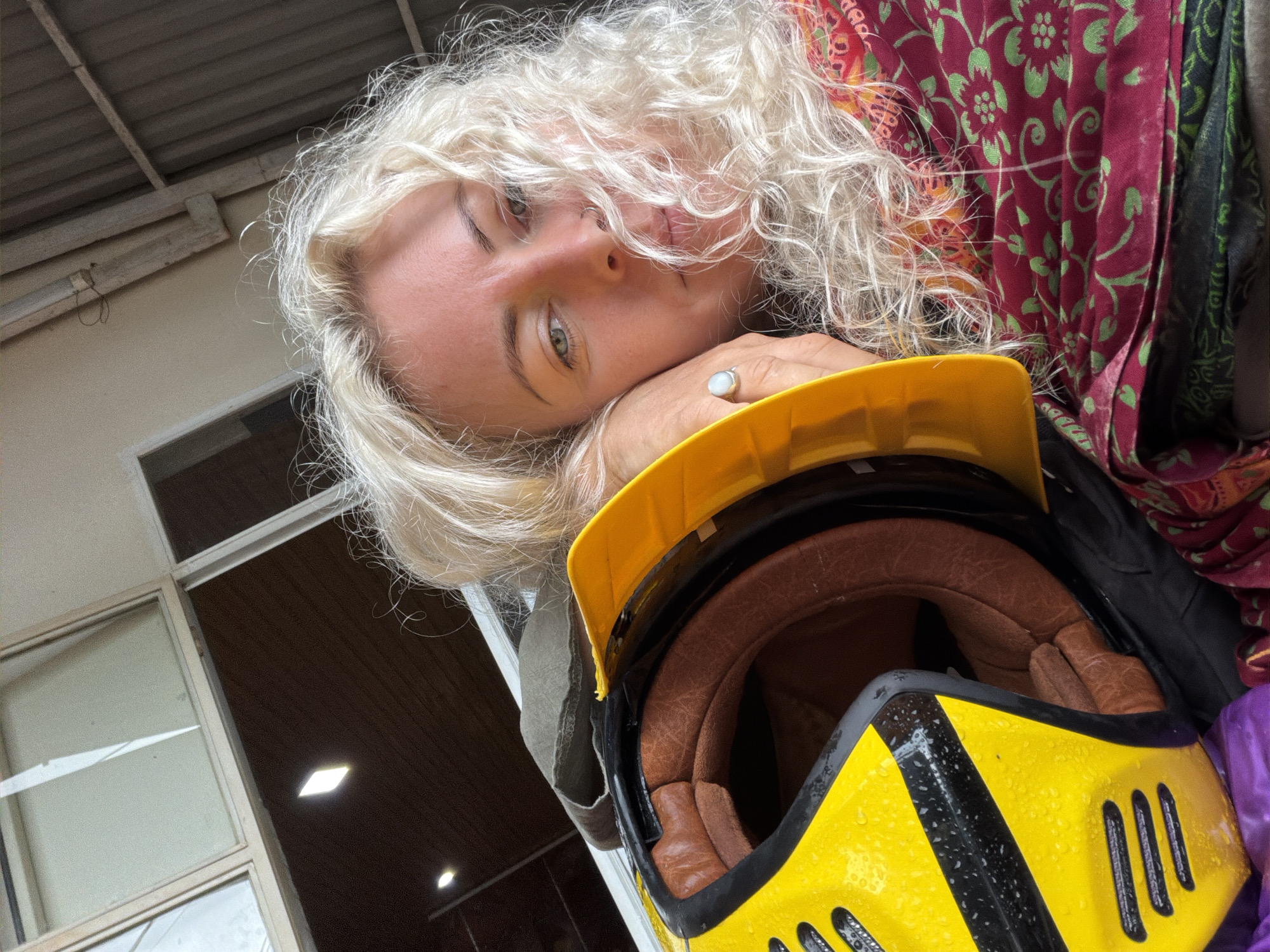 In Hướng Phùng Commune, Vietnam, a close-up shot captures a woman with blonde, curly hair resting her head on a yellow and black helmet. She is wearing a red and green patterned scarf. The background appears to be an open structure with a corrugated metal roof and some windows.