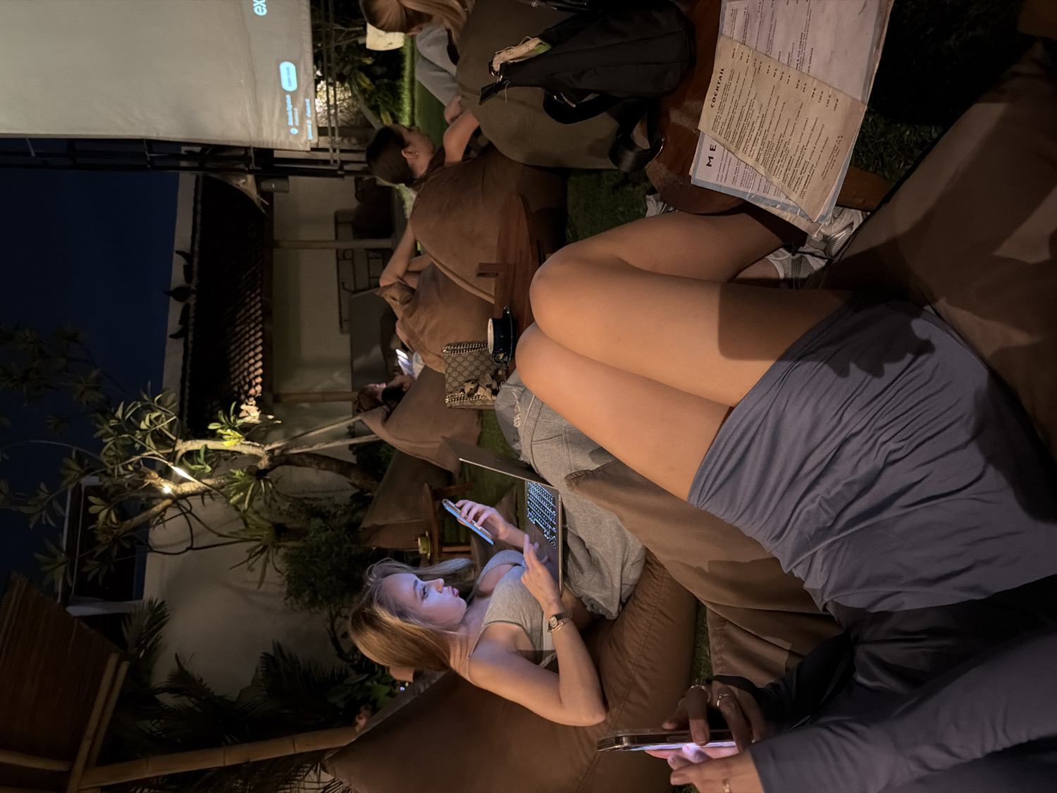 The media file captures an evening scene in an outdoor or semi-outdoor lounge area, likely a bar or restaurant, in North Kuta, Indonesia. Several people, primarily women, are relaxing on large, brown cushion-style seating.
In the foreground, a person is seen from the waist down, holding a smartphone with both hands. Beside them, another person is lying on their side, engaging with a laptop. This individual has bare legs and wears a blue top. Further back, a woman with long, blonde hair is lying on her side, illuminated by the glow of a smartphone or tablet she is holding. Other individuals are seated further in the background on similar cushions.
The setting includes lush tropical trees adorned with string lights, enhancing the evening ambiance. A large white projection screen is set up in the background, displaying white text against a dark interface, partially showing "explor..." and a UI toggle. A small table near the foreground seating holds paper menus; "COCKTAIL" is clearly visible on one, and "ME" (likely "MENU") on another. The time of day is clearly night or late evening, indicated by the dark sky and reliance on artificial light from screens and decorative string lights. The overall activity suggests a casual social gathering or entertainment event where people are relaxing and engaging with personal devices.