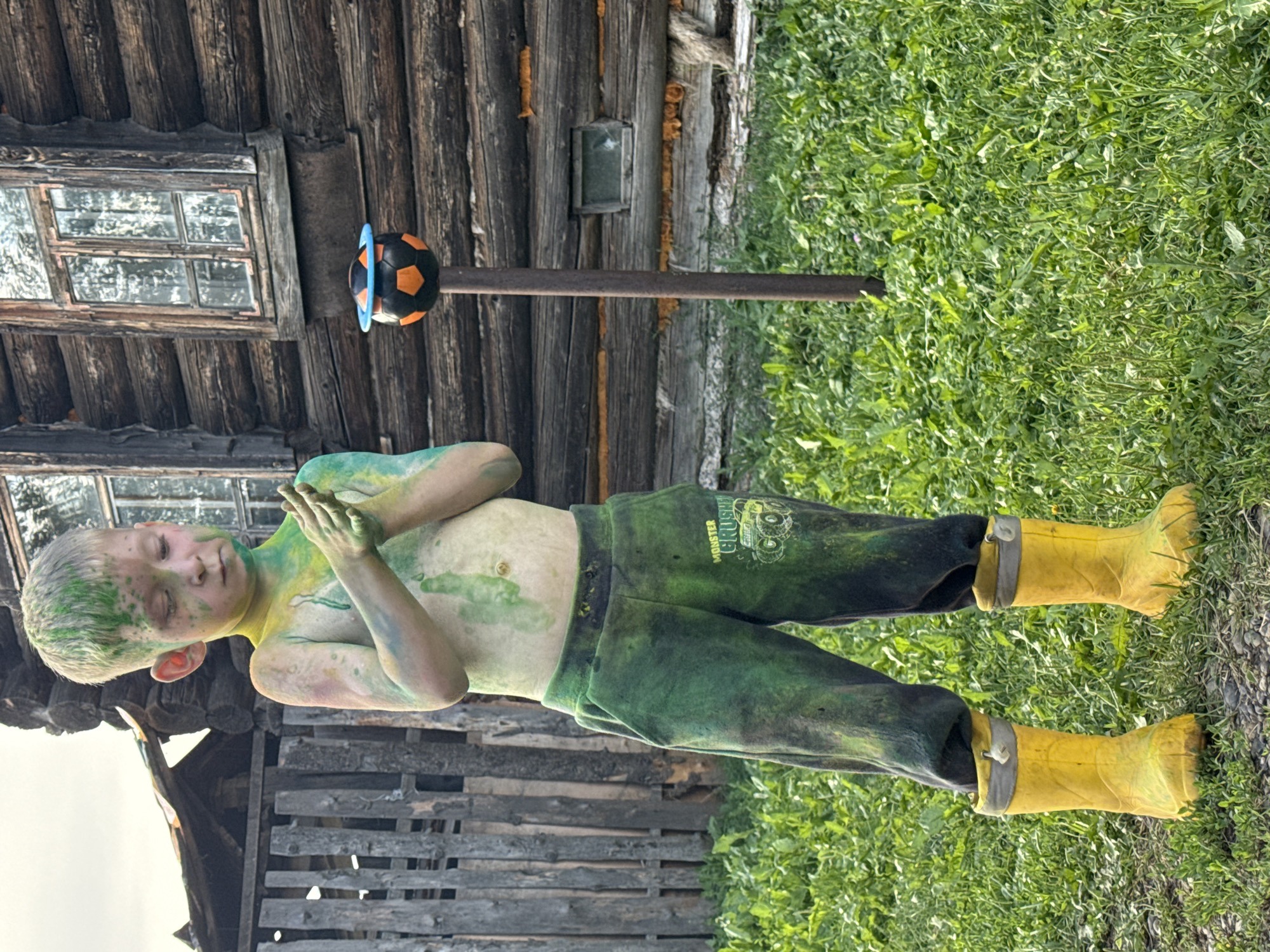 A young boy, covered in green and yellow paint, stands in front of a rustic wooden building. He is wearing dark green pants with a "MONSTER CRUSH" graphic and bright yellow rubber boots. The boy has his eyes closed and his hands clasped in front of his chest, suggesting a moment of contemplation or peace. He appears to be outdoors, surrounded by lush green grass and foliage. A metal pole extends from the ground behind him, supporting a toy soccer ball, half black and half orange, with a blue rim. The building behind the boy is constructed from weathered logs and has a small, multi-paned window. The scene suggests a playful or artistic activity, possibly related to a festival or creative project, taking place in a rural setting in Усть-Тунтор, Russia. The lighting suggests it is daytime, and the presence of green grass and leaves indicates a warmer season.