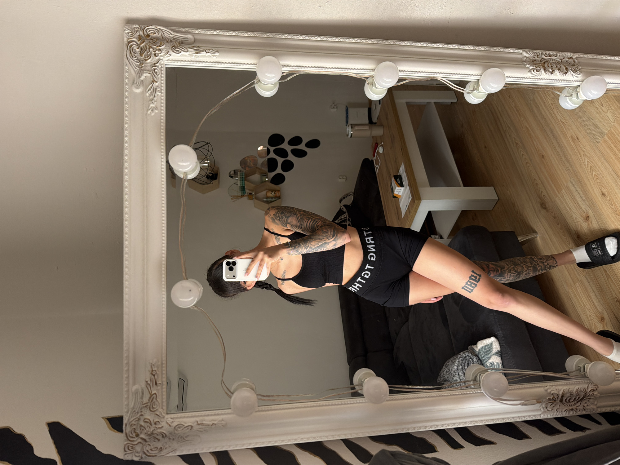 A woman with dark hair in braids and extensive tattoos on her arms and legs is taking a mirror selfie in a bedroom. She is wearing a black sports bra and black shorts. The mirror is large and ornate, with a white frame decorated with carved details and a string of round lights around its perimeter. In the reflection, a dark couch, a wooden coffee table with items on it, and a wall with hexagonal shelves and decorative circles can be seen. The floor is made of light-colored wood. The overall impression is of a well-lit, modern living space.