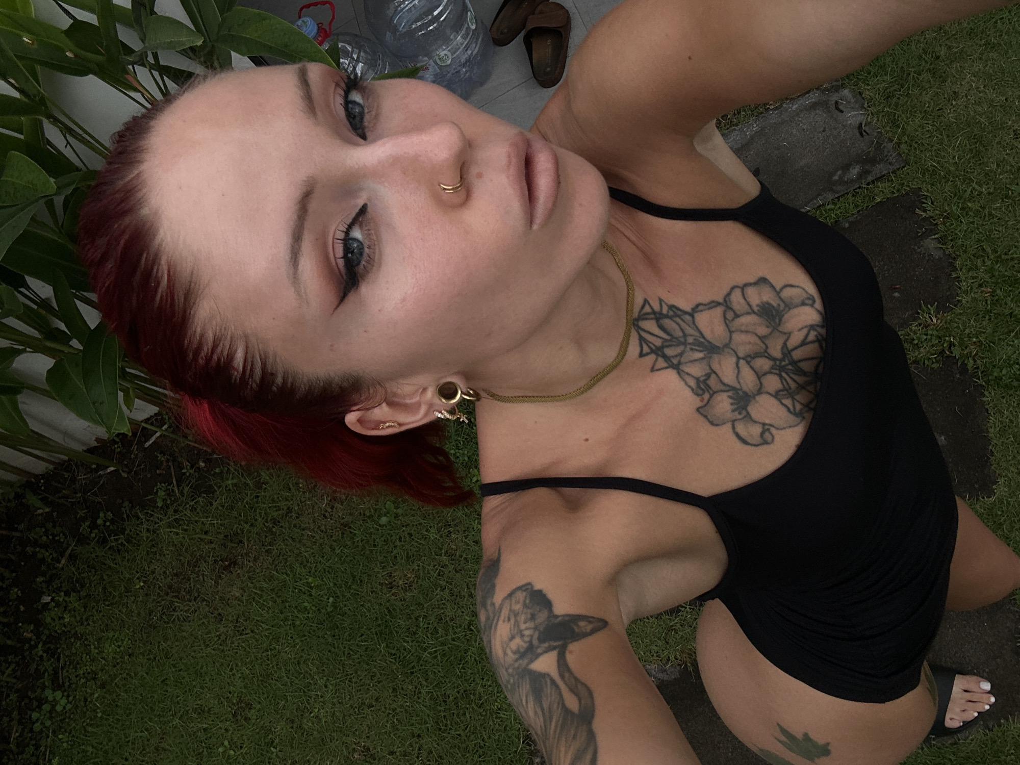A young woman with red hair pulled back is posing for a selfie outdoors. She is wearing a black one-piece swimsuit and has several tattoos on her left arm and chest. She is also wearing large hoop earrings and a nose ring. The background appears to be a grassy area with some plants and large water bottles. The scene is likely in North Kuta, Indonesia.