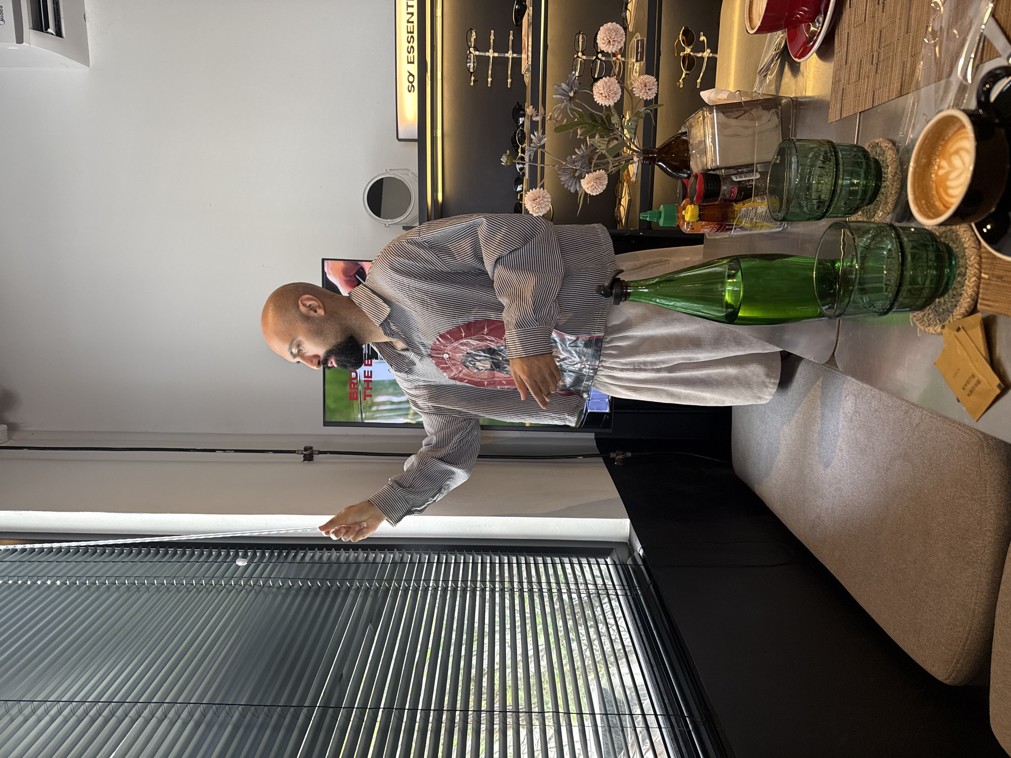 A bald, bearded man wearing a striped shirt and light-colored pants is standing indoors, drawing blinds on a large window. He is positioned near a table with several items, including two green glasses, a green bottle, condiments, and a coffee cup with latte art. In the background, shelves display sunglasses and other items, with a sign that reads "SO ESSENTIAL." The man appears to be in a shop or cafe. The overall lighting suggests it is daytime.