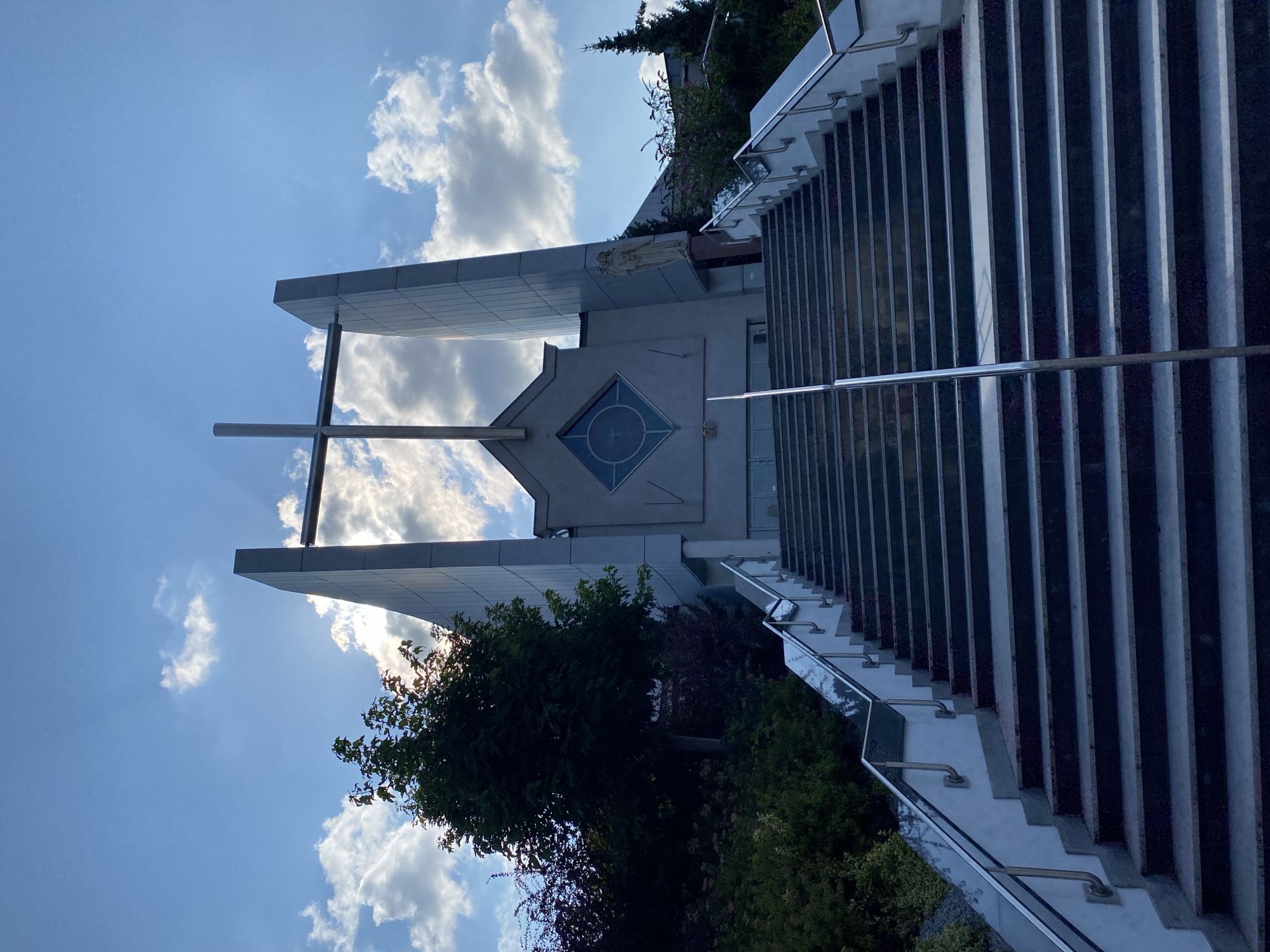 The image shows a wide, grand staircase leading up to a modern church building, likely located in the city of Przeźmierowo, Poland. The church features a prominent cross at its apex, silhouetted against a bright, partly cloudy sky. The architecture is characterized by angular, grey concrete structures. To the right of the entrance, a statue of a religious figure is visible. The staircase itself is made of stone with metal railings on either side. Lush greenery, including trees and bushes, borders the left side of the staircase, adding a touch of nature to the scene. The sun is shining through the clouds, casting rays of light that highlight the cross and the top portion of the church building. The overall impression is one of solemnity and architectural grandeur.