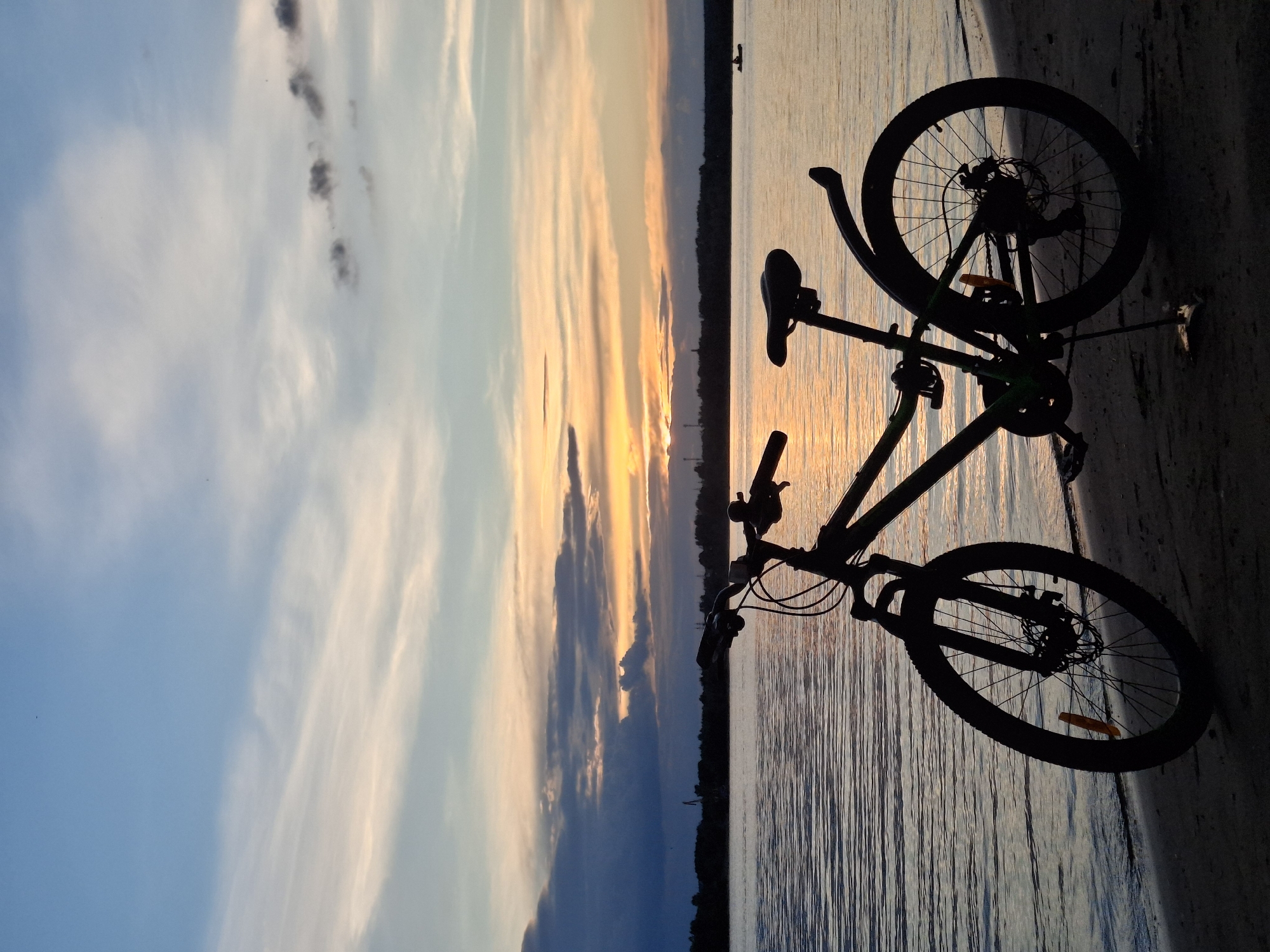 A silhouette of a bicycle stands on a sandy shore next to a wide body of water, likely the Kama River, in Chaikovsky, Russia. The sky above is a gradient of blue and orange, with wispy clouds indicating a sunset. The water reflects the colors of the sky. In the distance, a dark treeline is visible along the opposite bank, with a few industrial structures and a small boat with a person in it. The bicycle appears to be a mountain bike, with knobby tires and front suspension. It is leaning on its kickstand. The overall scene is tranquil and picturesque, capturing a moment of nature's beauty at dusk.