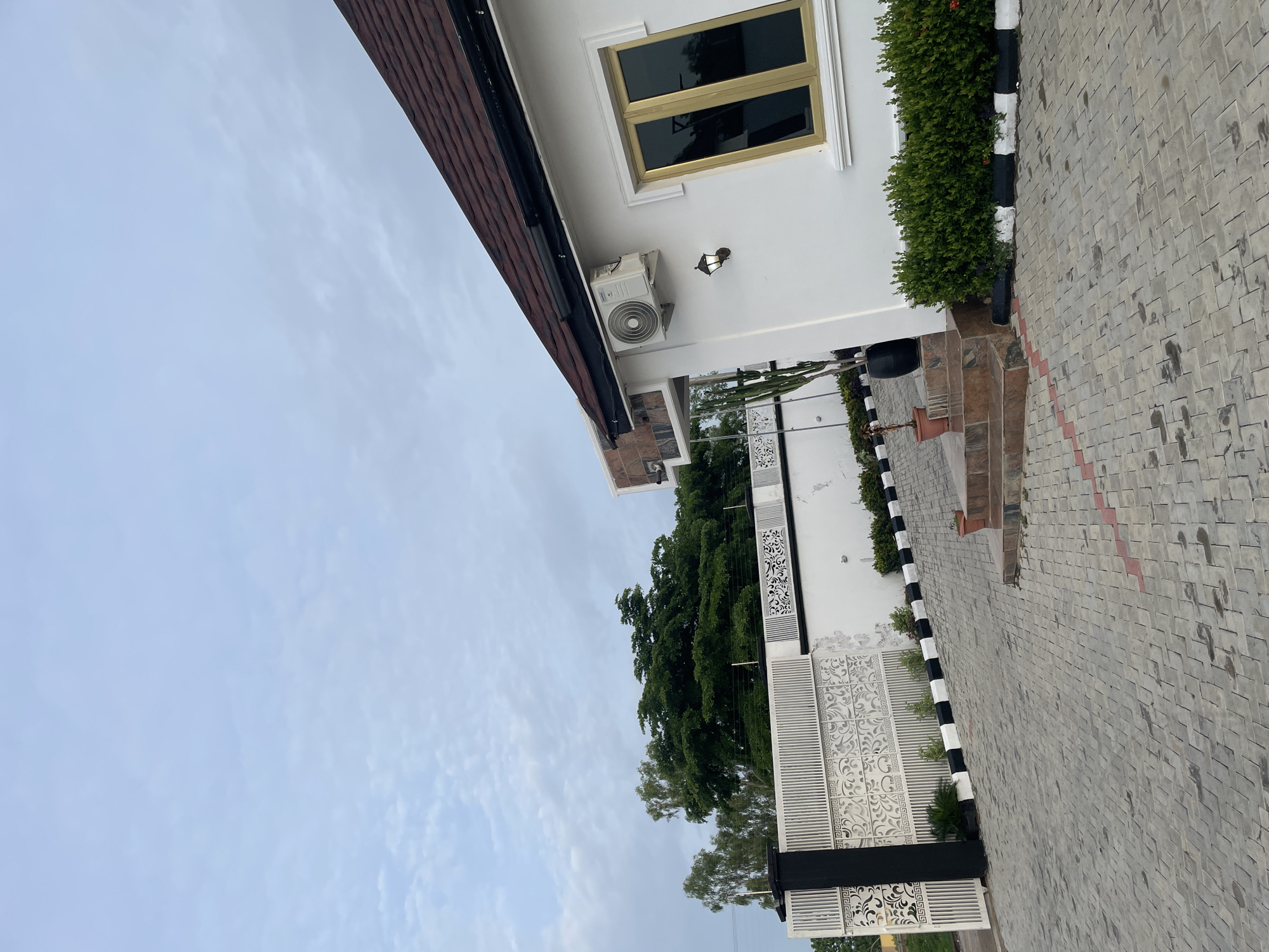 The image shows the exterior of a white building with a brown tiled roof in Jauro Yinu, Nigeria. The building has a gold-framed window and an air conditioning unit mounted on the exterior wall. A small outdoor lamp is also visible near the window. In front of the building, there is a paved driveway made of interlocking gray bricks with red and white markings. A low white wall with ornate black and white patterned metal fencing runs along the property line. Lush green bushes are planted along the edge of the driveway. There are a few potted plants, including a tall cactus in a black pot, placed on a small set of stairs leading to the building's entrance. The sky is overcast with scattered clouds. The overall scene depicts a well-maintained property with a modern architectural style.