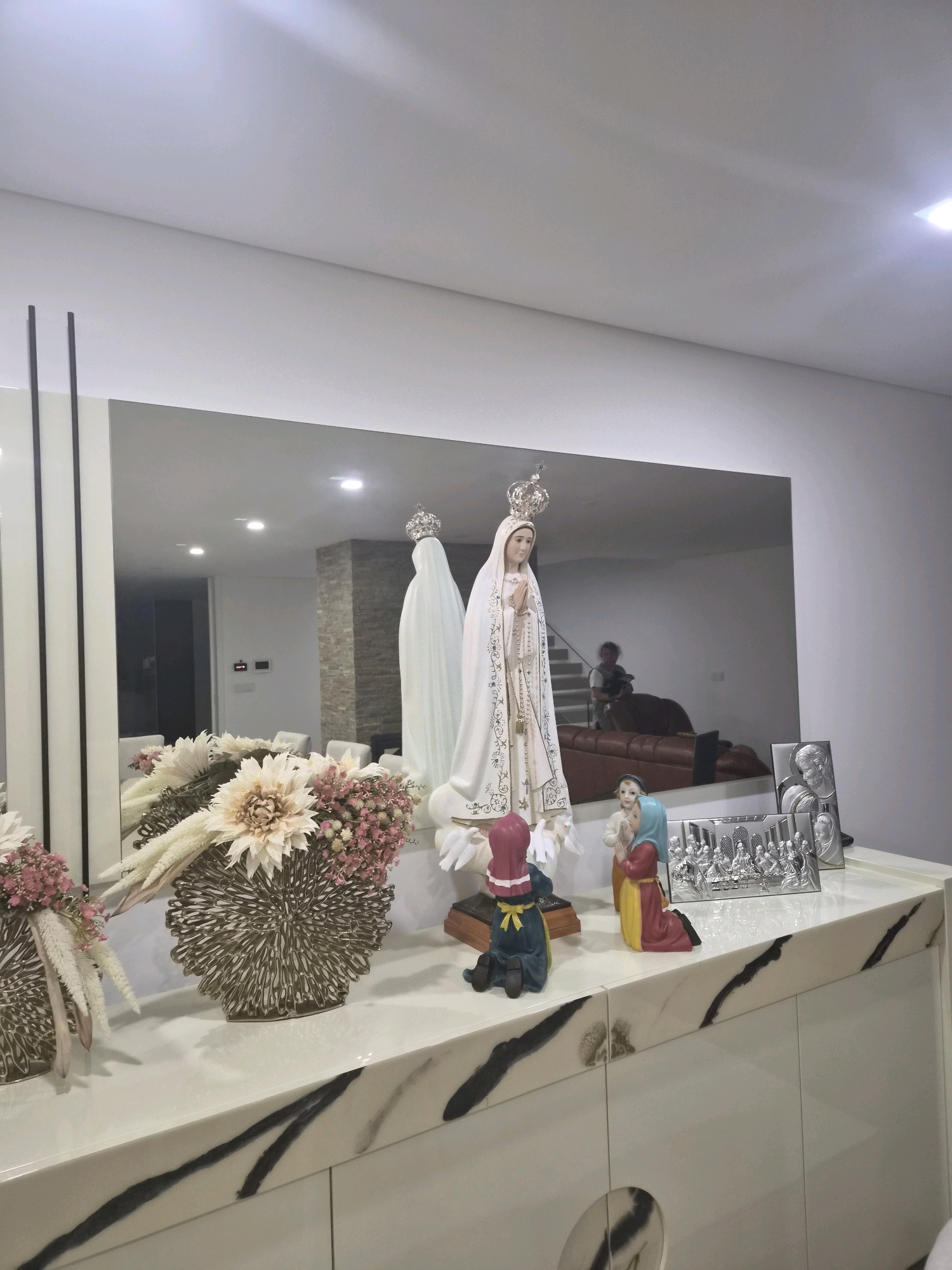 The image shows a decorative display on a white marble-topped cabinet. The display includes several religious figurines, including a statue of the Virgin Mary with a crown and rosary. There are also two smaller figurines of children in prayer, one wearing a blue headscarf and the other a red and blue habit. To the right of the figurines is a framed silver relief depicting the Last Supper. In front of the figurines are two ornate vases with dried flowers. A large mirror behind the display reflects the room, showing a staircase, a leather couch, and a person sitting on the couch. The setting appears to be an indoor living space. It is important to note that Barcelos, Portugal is known for its religious significance, particularly with the Sanctuary of Our Lady of Remedies, and this display may be representative of that devotion.