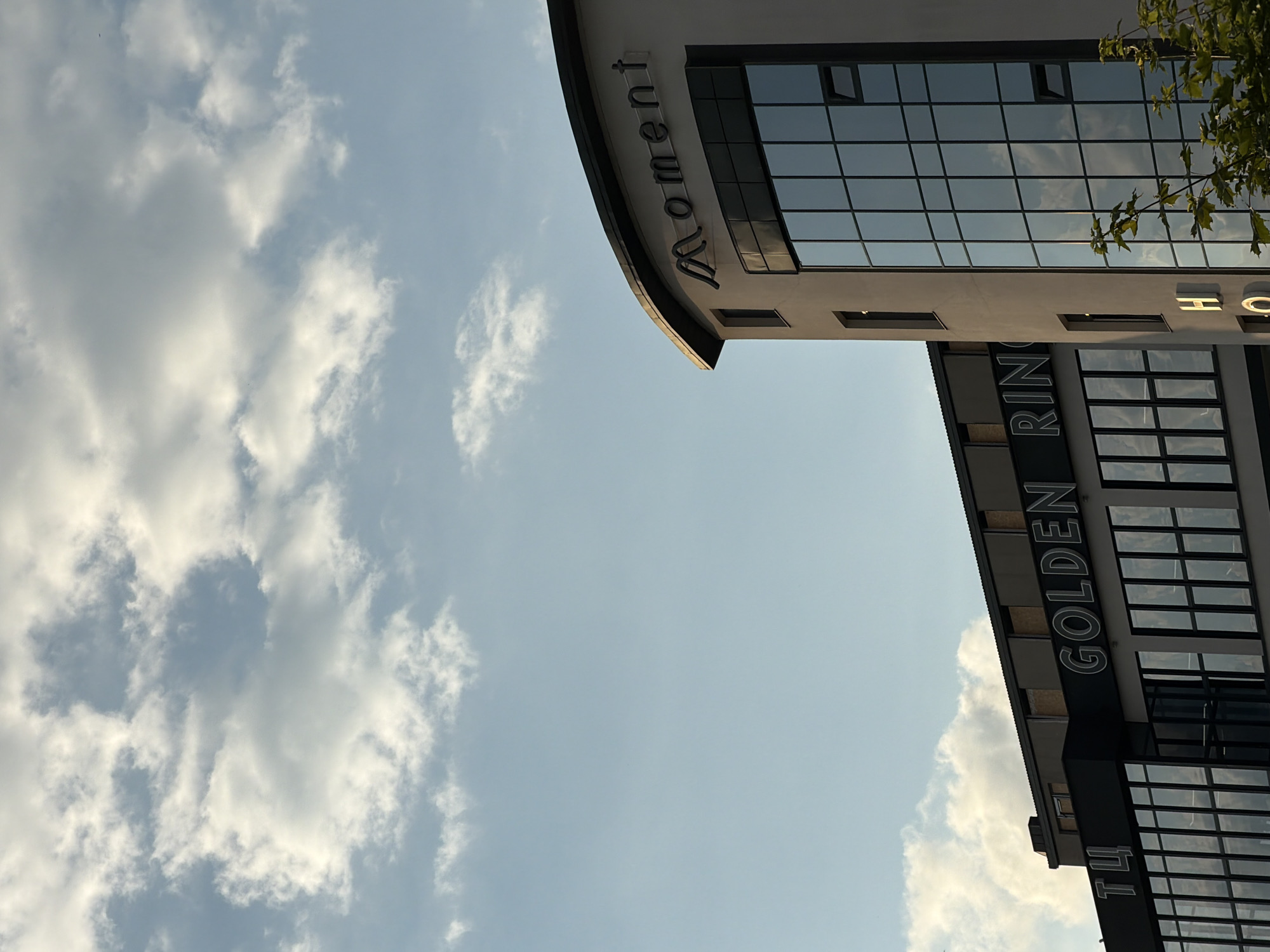 The image shows the facade of a modern building against a cloudy sky. The building has a curved section with the word "Moment" written in illuminated letters. Below this, a large glass facade reflects the sky. To the left, another section of the building is visible, with signage that reads "GOLDEN RING" and some Cyrillic letters, likely "ТУ". The building appears to be an office building or hotel. The scene is located in the city of Ternopil, Ukraine, indicated by the Cyrillic lettering on the building and the context provided. The lighting suggests it is either early morning or late afternoon. No people are visible in the image.