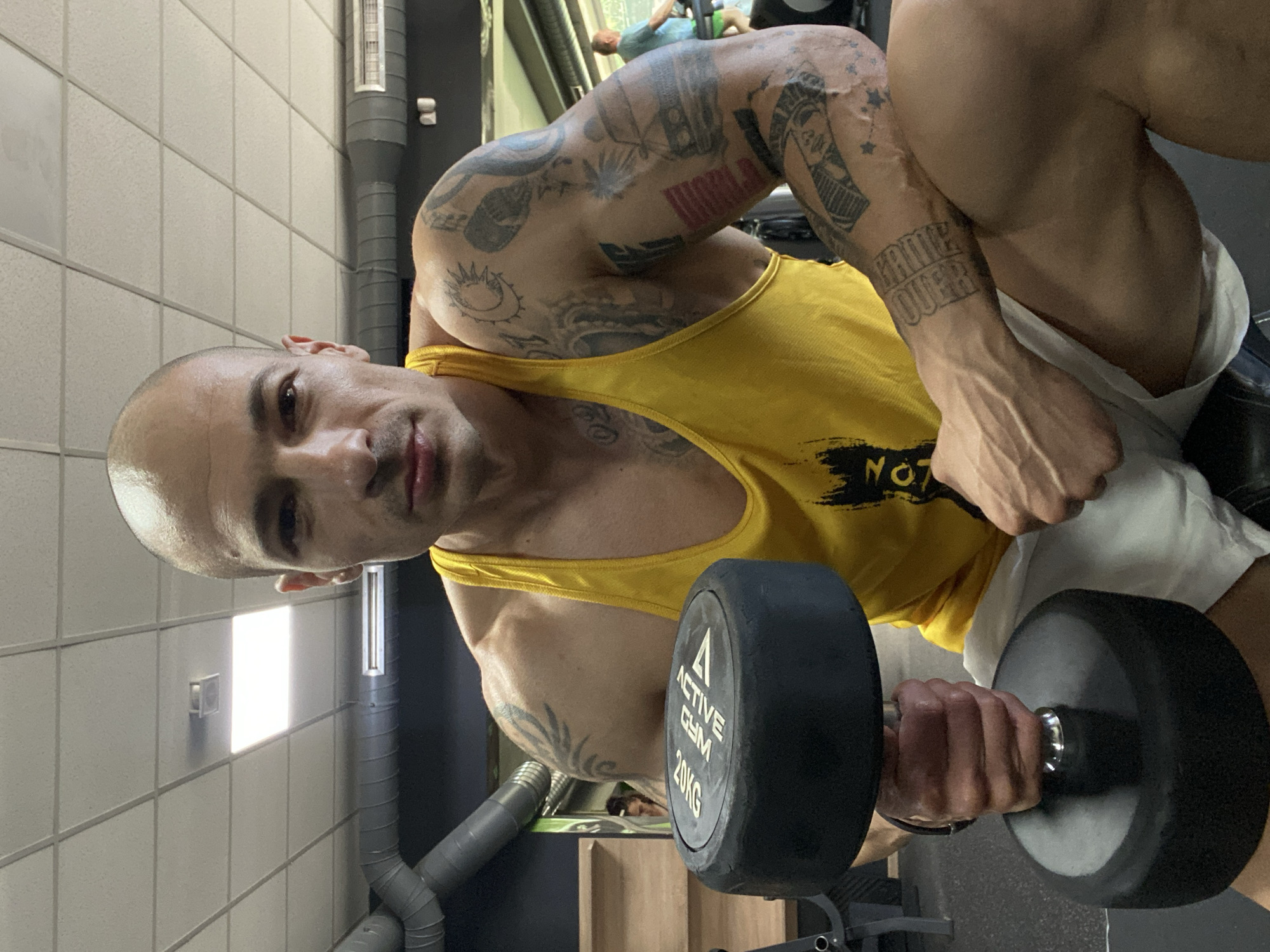 The image shows a muscular, bald man in a yellow tank top and white shorts lifting a 20kg dumbbell. He is heavily tattooed. The location appears to be a gym, identified by the "Active Gym" branding on the dumbbell, the weights, and the workout equipment. The gym is located in Pleven, Bulgaria.