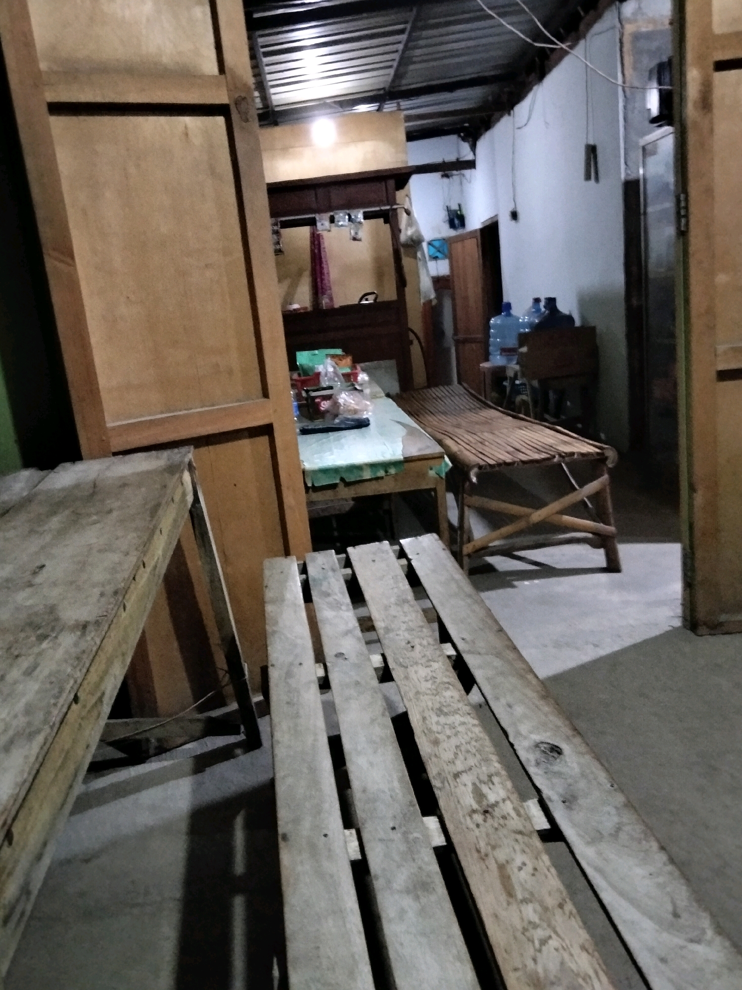 The image depicts an indoor scene, possibly a room or a storage area, in Mrican, Indonesia. The environment is cluttered and appears to be in a state of disarray. In the foreground, there are two wooden structures that resemble benches or tables, constructed from planks of wood. One is on the left, angled diagonally, and the other is in the center, closer to the viewer and running horizontally.
The background reveals more of the interior, including what seems to be a table with items on it, such as plastic bottles and possibly food packaging. A portion of a door or a large wooden partition is visible on the left side of the frame. Further in the background, there is a wall with some hanging objects, and the ceiling appears to be made of corrugated metal. Two large blue water cooler bottles are placed on a stand. The lighting is dim, with a single light source visible overhead, creating shadows on the floor and walls. The overall impression is of a functional but unkempt space.