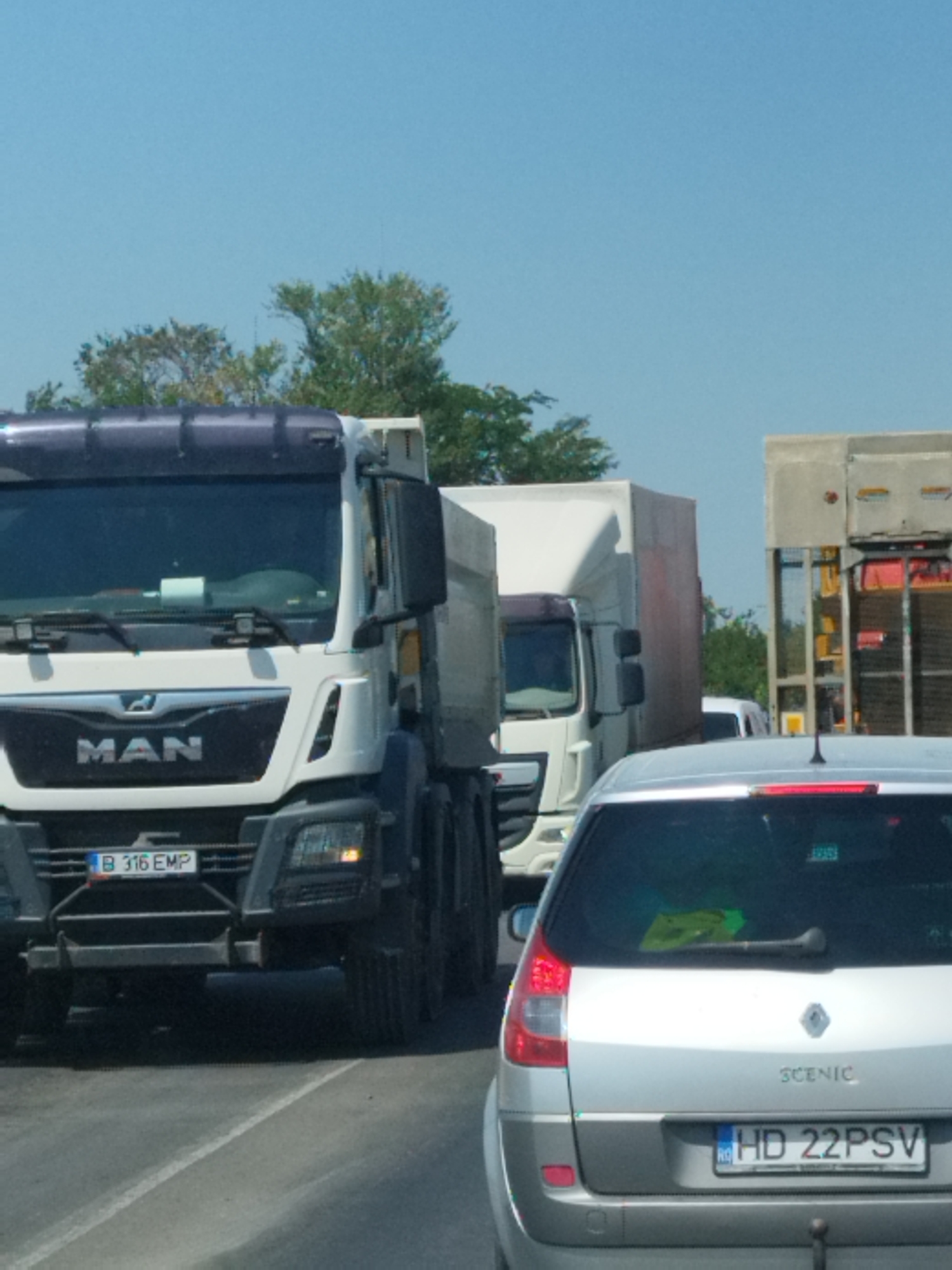 The image depicts a traffic scene on a sunny day, likely in or near Clinceni, Romania. In the foreground, a silver Renault Scenic is parked. Behind it, a white truck with the "MAN" logo on its grille is visible. Further back, another white truck with a similar trailer is in the lane. The sky is clear and blue. The road appears to be asphalt with a white lane marking. The overall impression is one of a traffic jam or slow-moving traffic on a highway or main road.