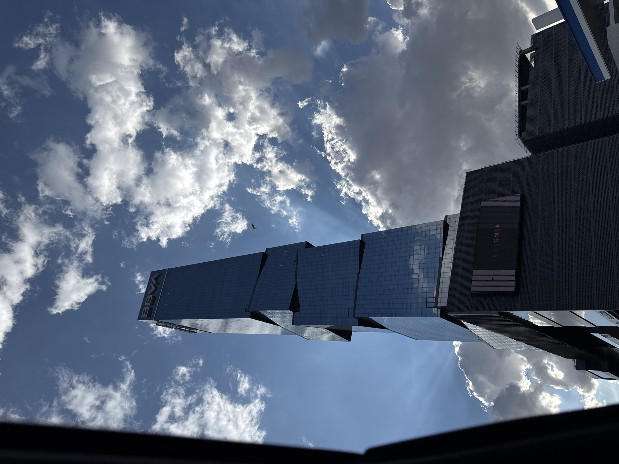 The image presents a low-angle view of modern buildings against a sky filled with cumulus clouds, taken in San Bernardino Tlaxcalancingo, Mexico. The building on the left has "BBVA" printed at the top of the building, and the building on the right is a black modern building. The angle suggests the photograph was taken from a low vantage point, possibly inside a building or from a vehicle. The sky includes a mixture of blue patches and white clouds, with some areas appearing darker, possibly due to shadows or thicker cloud cover. The buildings feature a modern architectural design, with multiple sections appearing to step back, creating a layered effect. The building to the right has a sign "IGNIA" on the side. There is a bird flying in the sky.