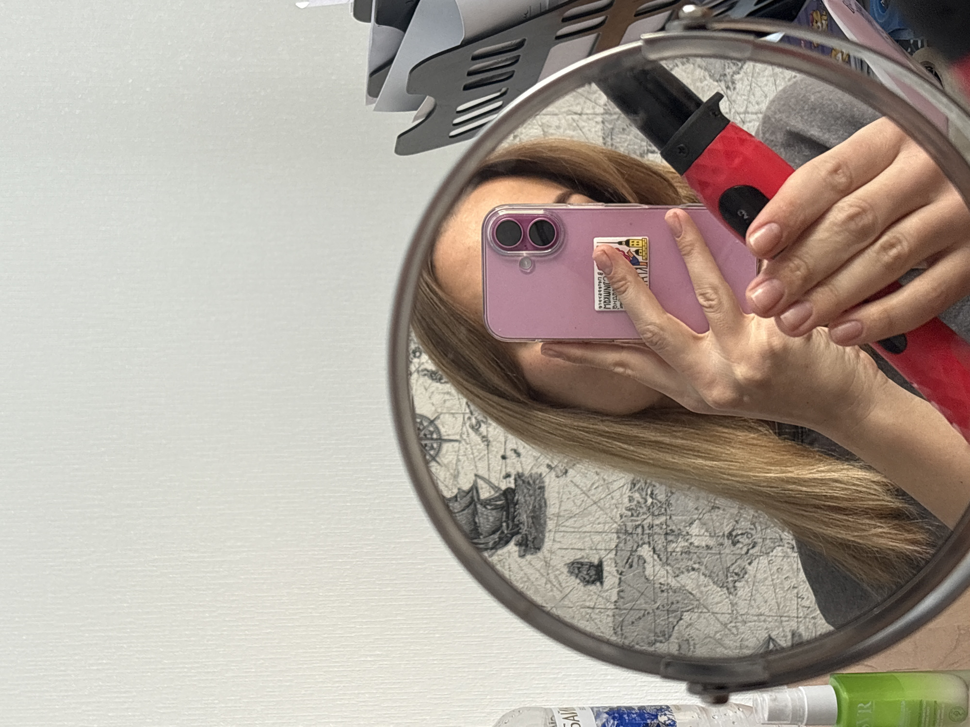 A person is captured in a mirror selfie, reflecting the upper part of their body, head, and hands in a round, metallic-framed mirror. Their long, light brown hair cascades around their face, which is mostly obscured by a pink/purple smartphone held in a clear case. The phone displays prominent dual camera lenses and features a sticker on its back with an illustration of buildings and visible text, including "DUNNKOW" and Cyrillic characters that appear to be "РУНКОВА" or "РУННОВА".
The person’s right hand holds a red and black hair styling tool, possibly a curling iron or straightener, with an "ON/OFF" control visible. Their fingernails are neatly manicured with a clear or natural polish. They are dressed in what appears to be a grey top.
The background reflected within the mirror is a light-colored wallpaper depicting an old-world map, complete with a compass rose, stylized ship illustrations, and outlines of landmasses. Outside the mirror's reflection, a white, textured wall serves as the primary background. Above the mirror, dark-colored kitchen utensils, such as spatulas, are neatly organized on a rack, indicating a domestic setting.
At the bottom edge of the mirror, two bottles are visible: one blue and white with partial Cyrillic text "БАЙ" (possibly from "Байкал"), and another green and white bottle labeled "SR." This indoor scene, likely a routine moment of getting ready, is set in Ust-Ilimsk, Russia.