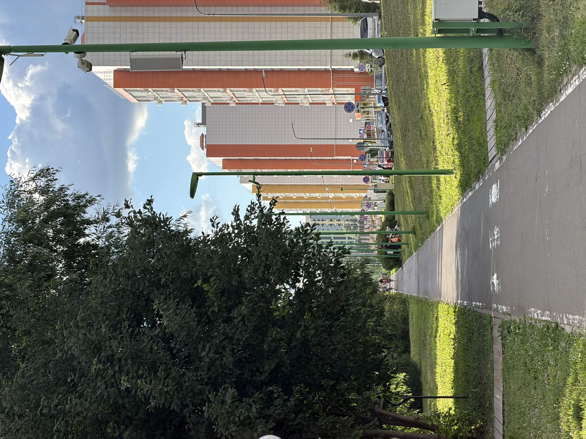 The image shows a paved pathway running through a grassy area with trees on the left and modern apartment buildings in the background on the right. The pathway has white markings and a white border. Several people are walking or standing on the pathway and in the grassy areas. The apartment buildings are tall and feature orange and white facades, with visible windows and balconies. Green lampposts are placed along the pathway. Cars are parked on the street in front of the buildings. The sky is blue with scattered white clouds. The scene appears to be a residential area in a city.