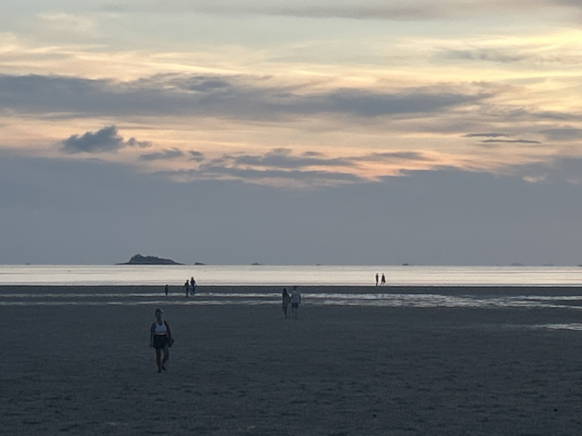 The image shows a beach scene in Baan Sri Thanu, Thailand, at dusk. The sky is filled with layers of clouds, tinged with pale orange hues from the setting sun. The beach is wide and flat, with people walking across it. In the foreground, a woman in athletic wear walks towards the viewer. Further away, a couple walks together, and a small group is gathered near the water's edge. In the distance, a small island is visible on the horizon.