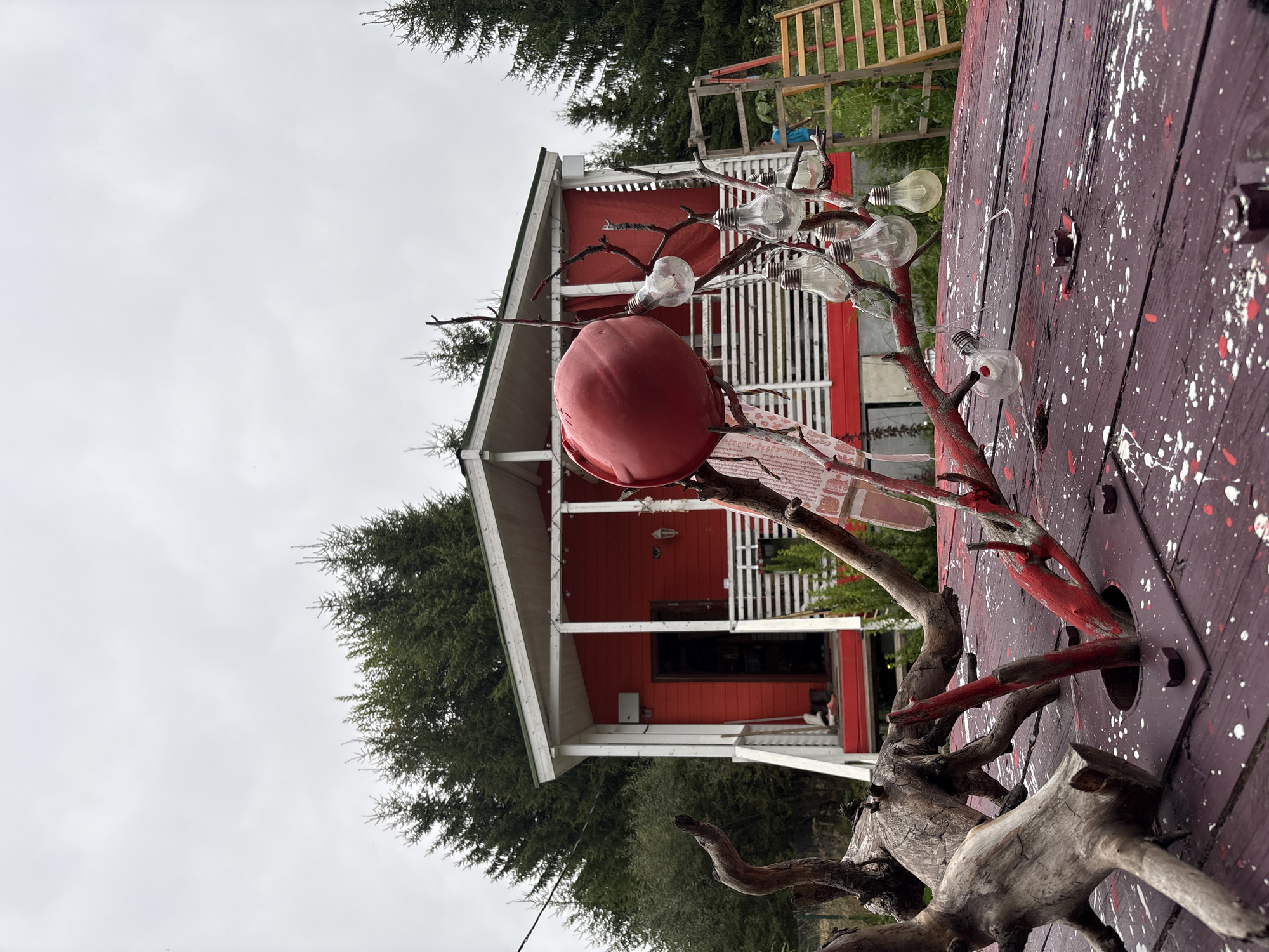 The image depicts an outdoor scene in front of a red house. In the foreground, a weathered wooden table is visible, painted dark red with white and red splotches. Several pieces of gnarled driftwood are embedded in or resting on the table, with one prominent piece having a red-painted branch adorned with clear light bulbs. Behind the table, the red house has a white-trimmed porch with a white railing. A red tarp is visible behind the railing on the porch. The sky is overcast and a row of dark green evergreen trees forms the background. To the right of the house, a wooden ladder leans against an unseen structure. The overall impression is of a rustic, somewhat eclectic outdoor setting. The specific location of Lyady, Russia, is not visually ascertainable from the image itself.