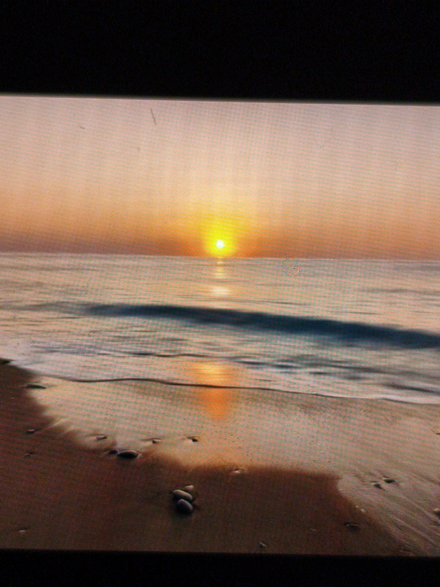 The image shows a beach at sunset. The sun is centered on the horizon, reflecting on the calm water. The foreground is the wet sand of the beach with scattered pebbles. The sky is a gradient of orange and yellow.
There is no indication of Devin, Bulgaria.