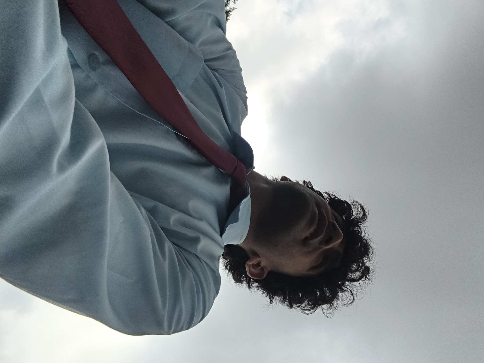 A low-angle shot captures a young man with curly dark hair, wearing a light blue collared shirt and a maroon tie. He is looking upwards against a cloudy, overcast sky. The image appears to be taken outdoors. The man's face is partially obscured by his upward gaze, but his features are visible. His left arm is bent and positioned in front of his chest, with his hand out of frame. The fabric of his shirt is wrinkled, suggesting movement or a relaxed posture. The background consists entirely of the sky, which is a uniform grey with some lighter areas indicating clouds. No specific landmarks or city features are discernible, making it impossible to pinpoint a location within Dharamshala, India, or any other city. The scene is simple and focused on the individual and the sky.