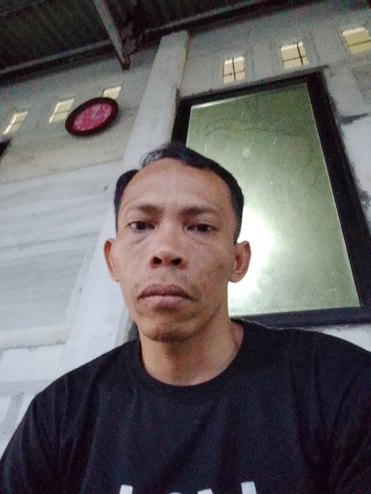 The image is a selfie taken indoors in Bukit Ketok, Indonesia. A man with short dark hair and a black t-shirt is the primary subject. Behind him is a wall with small, square windows near the ceiling and a framed window. A red-framed clock hangs on the wall to the left of the window. The lighting appears dim and possibly artificial.