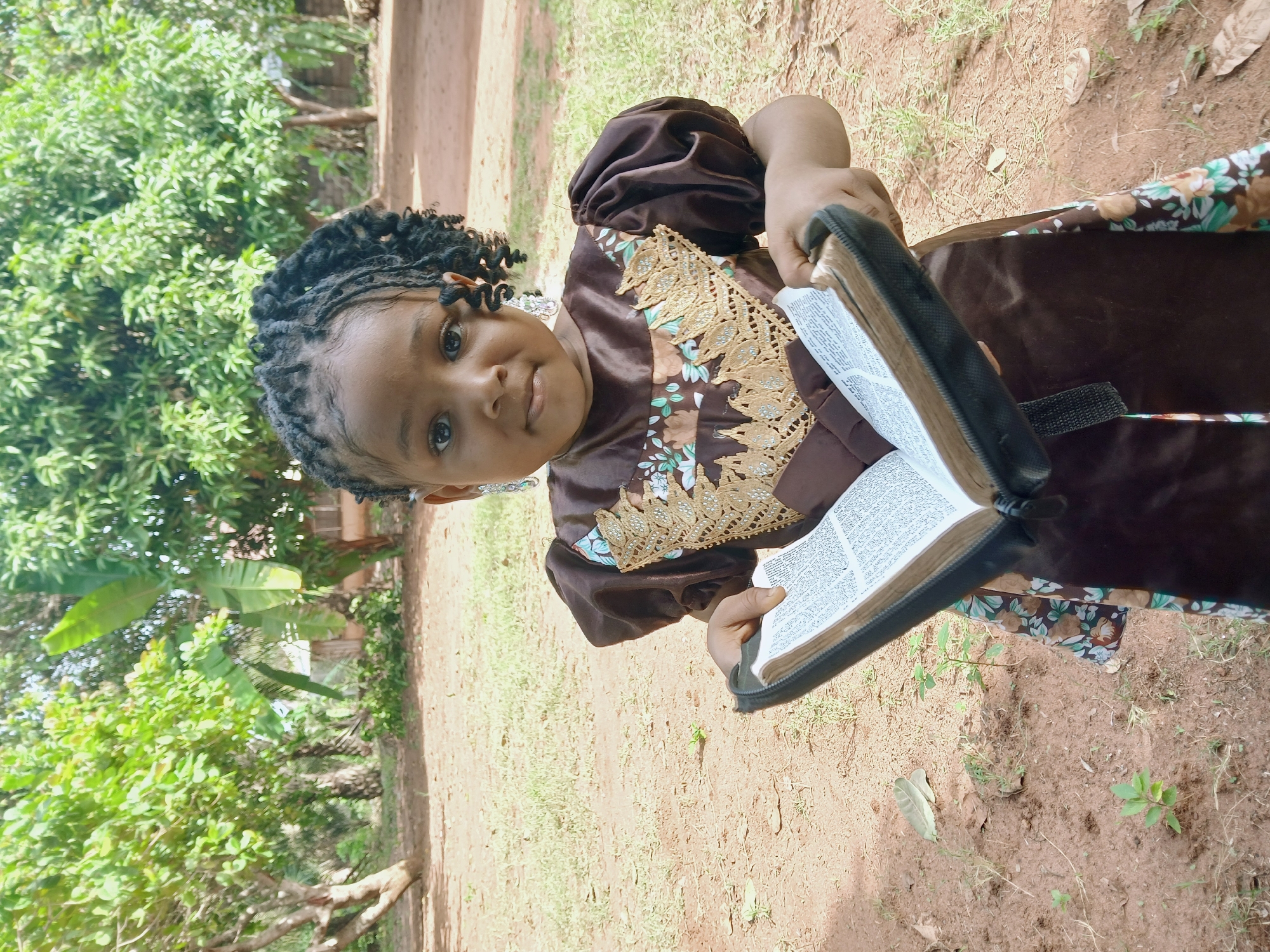 A young girl is standing in a dirt clearing, holding a book with visible text, likely a Bible, open. She is wearing a brown dress with floral accents and a gold-colored lace-like embellishment on the chest. The girl has her hair in braided or twisted strands. The background includes green trees and foliage. The location is likely a rural or semi-rural area in Ankpa, Nigeria, based on the landscape and vegetation.