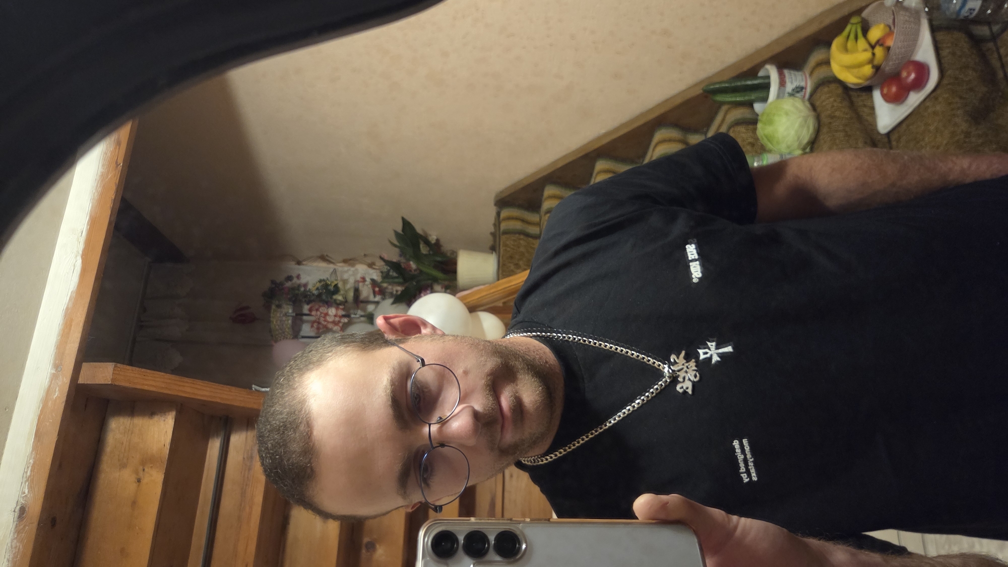 The image is a slightly distorted selfie taken in what appears to be a home setting. The subject is a light-skinned male with short brown hair, a light beard, and is wearing round, thin-rimmed glasses. He is also wearing a black t-shirt with a silver chain and a cross pendant. The t-shirt has some text on it. On the left side of the shirt, near the collar, are the words "212 KINGS". Below the pendant, also on the shirt, is text written in what appears to be Cyrillic script, but it is reversed and hard to read.
The background of the image shows a staircase with wooden steps and a railing. To the right of the stairs, on a shelf, is a basket of fruit including bananas, apples, and possibly a peach. There is also a head of lettuce and a cucumber visible. Further in the background, there is a white candle and some flowers in a vase. Several white balloons are also visible, suggesting a possible celebration. The overall lighting suggests it is daytime, or at least a well-lit interior. The reflection in the mirror suggests a cluttered but lived-in environment. There are no overt indications of weather, but the indoor setting with balloons might hint at a festive occasion. The subject's expression is neutral. The image quality is fair, with some graininess.