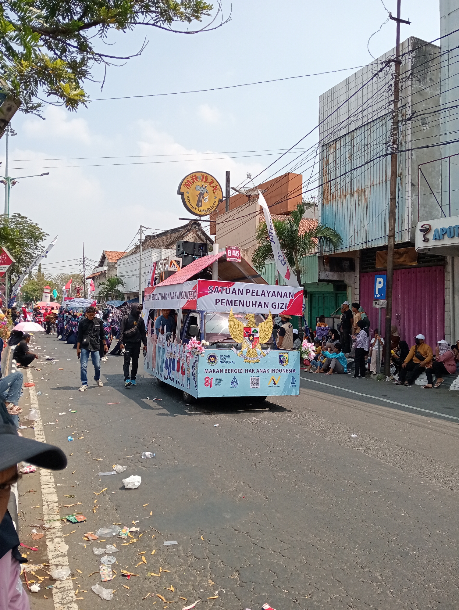 This is a medium shot, eye-level, outdoor photograph taken on a sunny day in the city of Demak, Indonesia. The image captures a street parade with a decorative float as the central subject. The float, a light blue truck adorned with banners and graphics, features the Garuda Pancasila emblem. Banners on the float read "SATUAN PELAYANAN PEMENUHAN GIZI" and "MAKAN BERGIZI HAK ANAK INDONESIA" with an "81" numeral prominently displayed. A group of people, possibly participants in the parade, are walking alongside and on the float, some in matching uniforms.

The street is paved and appears somewhat worn, with some cracks visible. Litter, including plastic bags, wrappers, and fallen leaves, is scattered on the road and sidewalk. On the right side of the street, several buildings are visible. One building has a light blue corrugated metal facade and appears to be a shop with the word "APOT" partially visible, indicating it might be a pharmacy. A blue parking sign with a white 'P' is also visible on the street.

On the left side of the street, more people are gathered, watching the parade. Some are seated on the curb, while others stand. A large tree with green foliage is in the upper left corner of the frame, casting some shadows. Power lines and streetlights are also present, indicating an urban environment. The sky is clear and blue. The overall atmosphere suggests a festive community event.